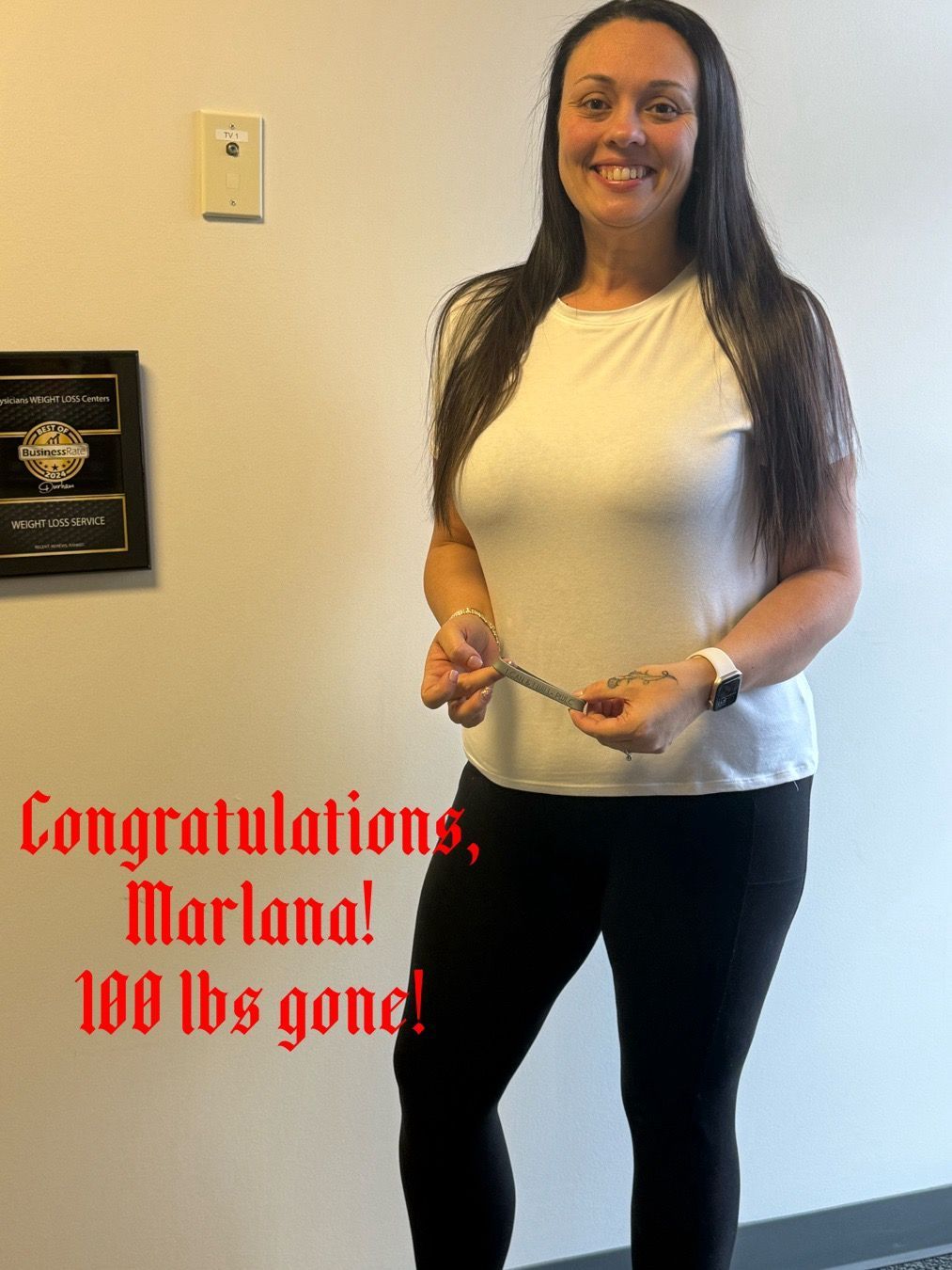 Woman smiling, holding a measuring tape. Text: Congratulations, Marlana! 100 lbs gone! In a hallway.