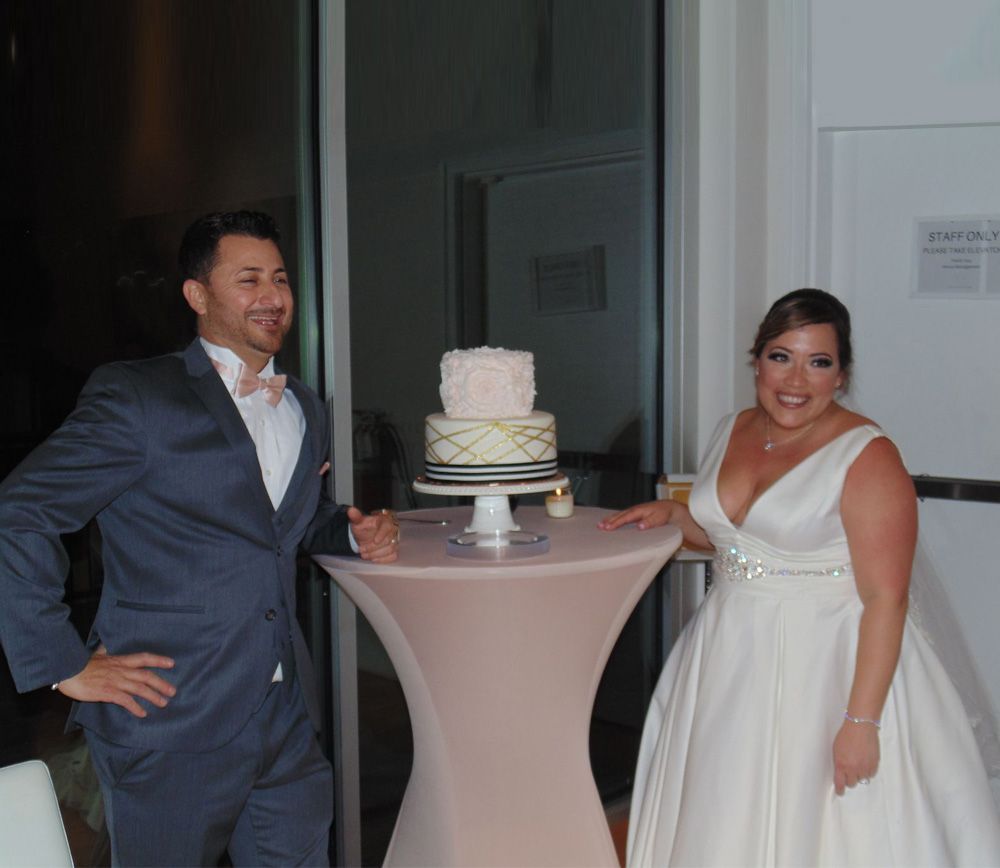 Bride and groom next to a tiered cake on a table. The groom wears a blue suit, the bride a white dress.