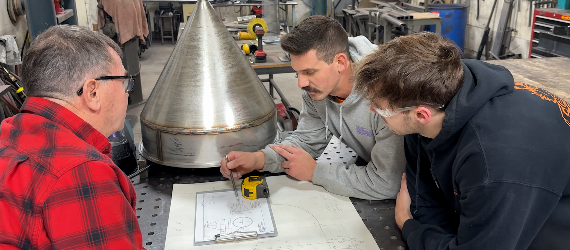 Three people examine a drawing near a metal cone in a workshop. A person measures with a tape measure.