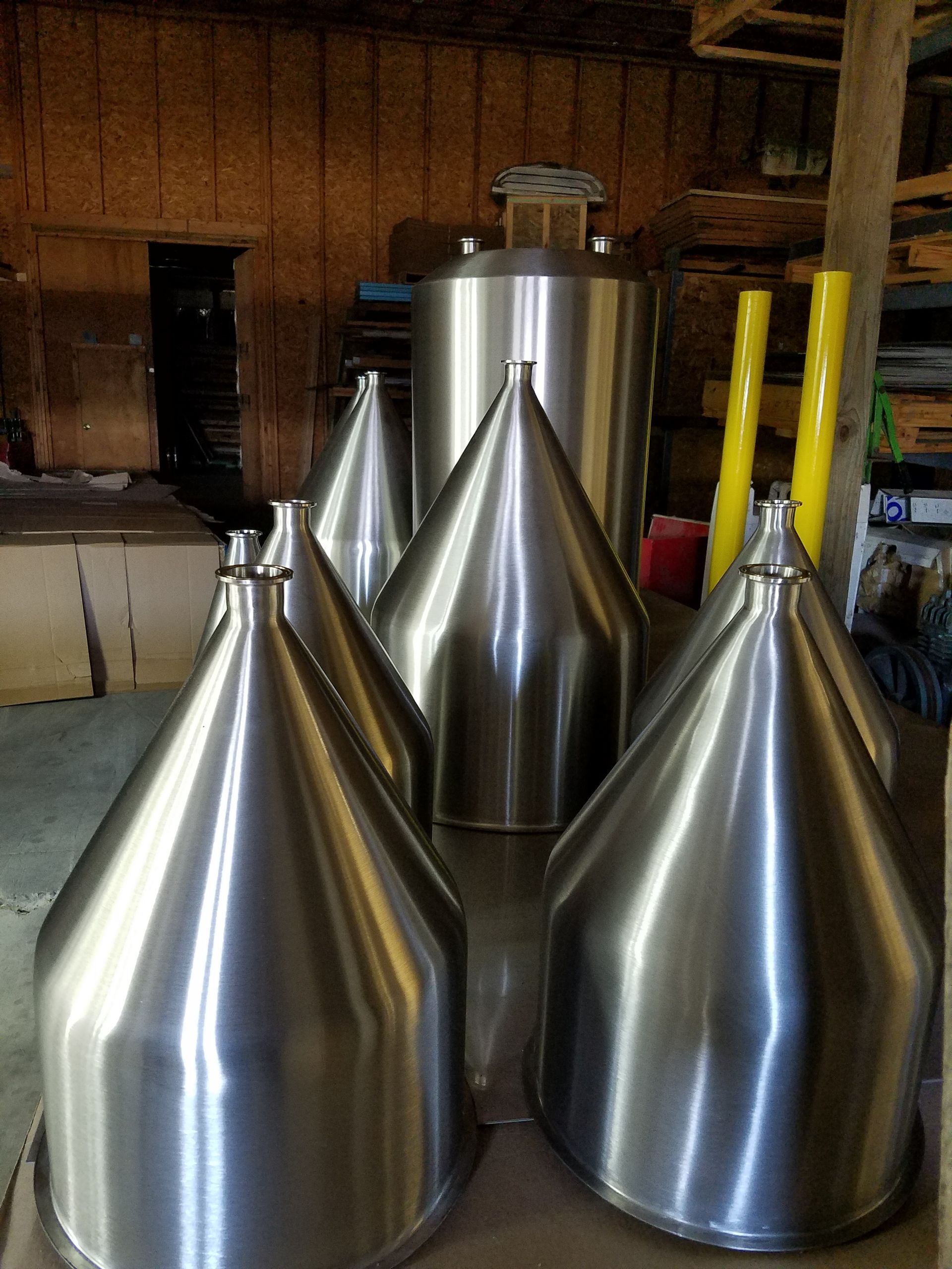 Shiny, conical stainless steel tanks in a workshop setting with a cylindrical tank in the background.