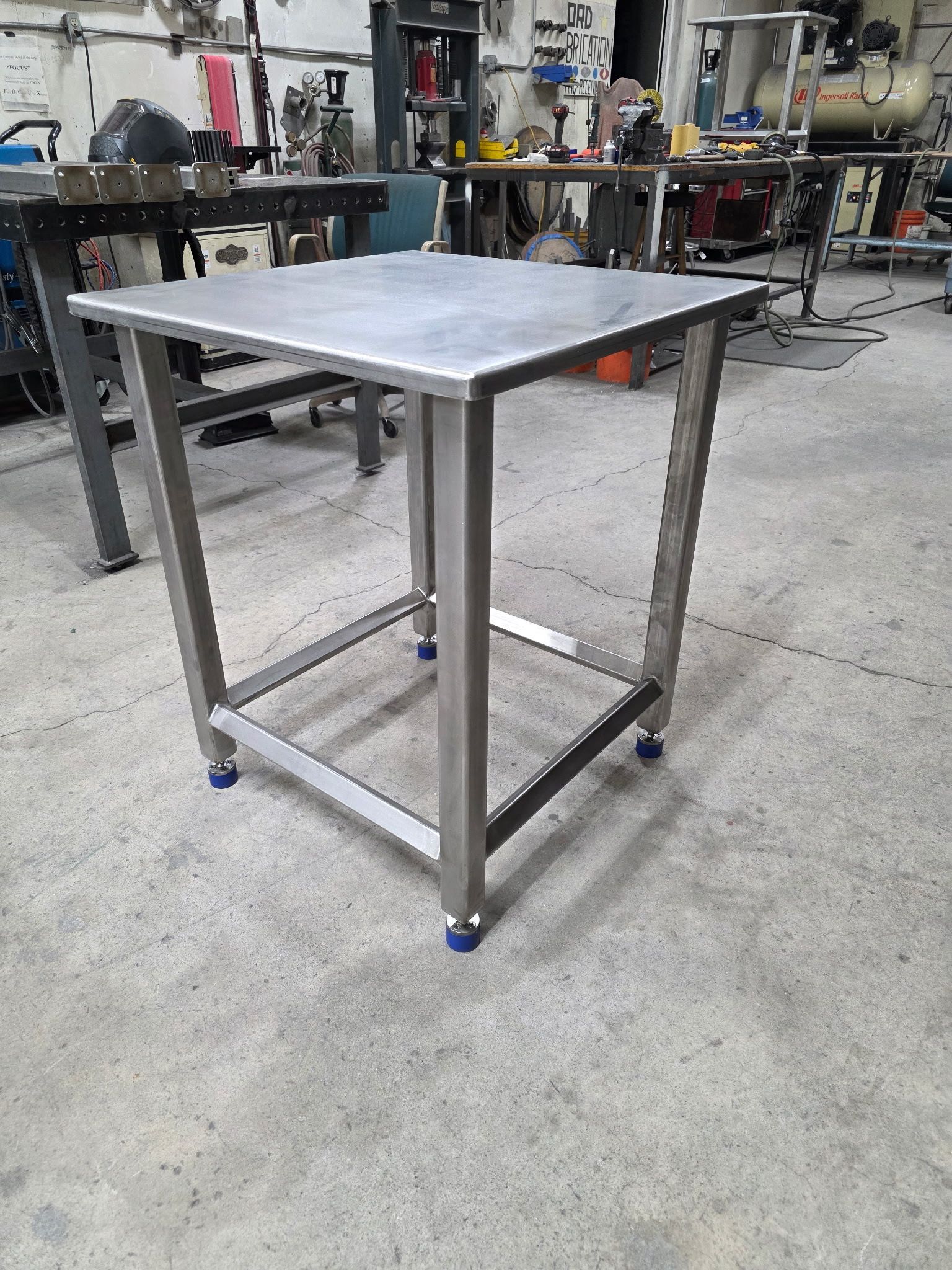 Stainless steel square work table on a concrete floor with blue capped legs in a workshop.