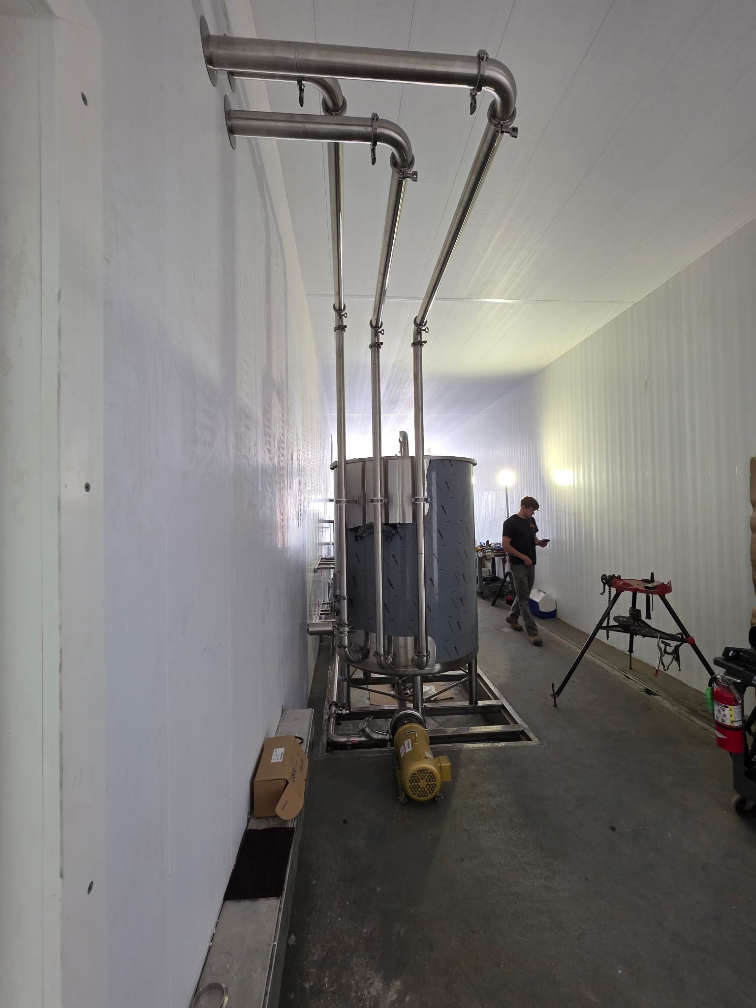 Industrial piping and tank mounted on a wall in a white-walled room; a person works in the background.