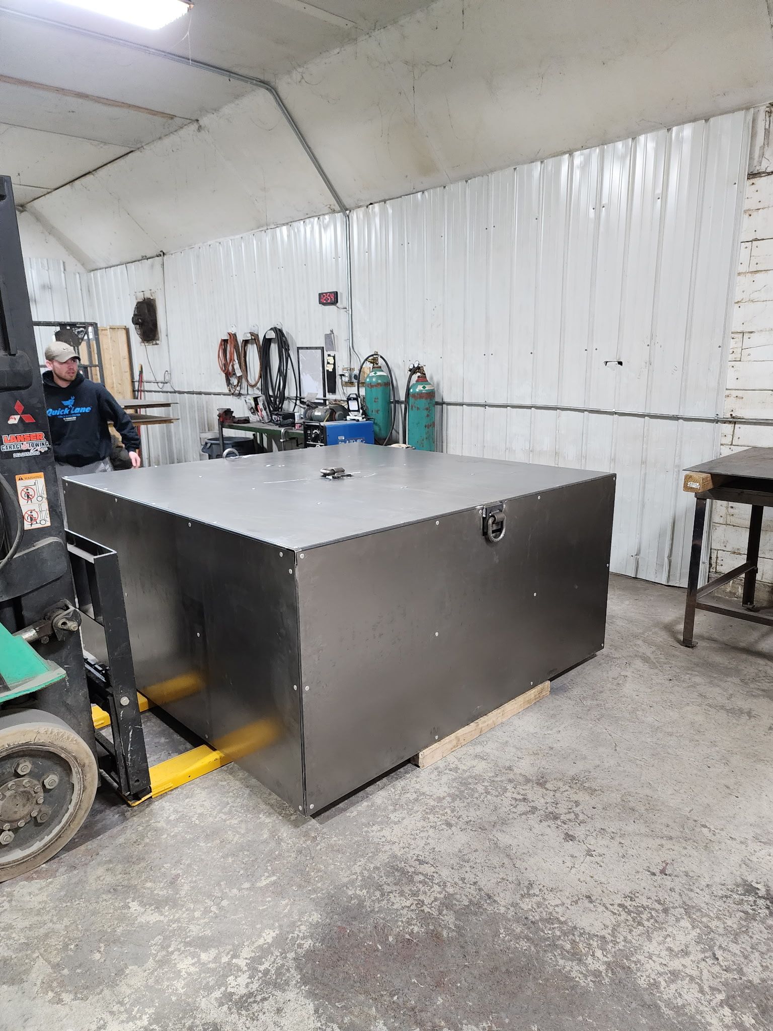 A large, metal box being moved by a forklift in a workshop. A person is nearby.