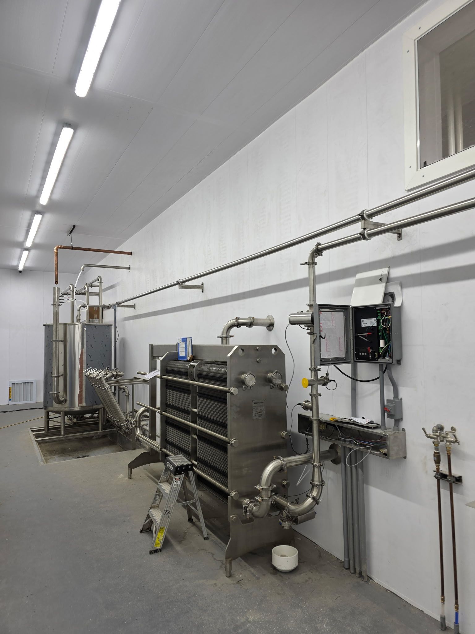 Industrial equipment in a white-walled room with stainless steel pipes and tanks for processing.