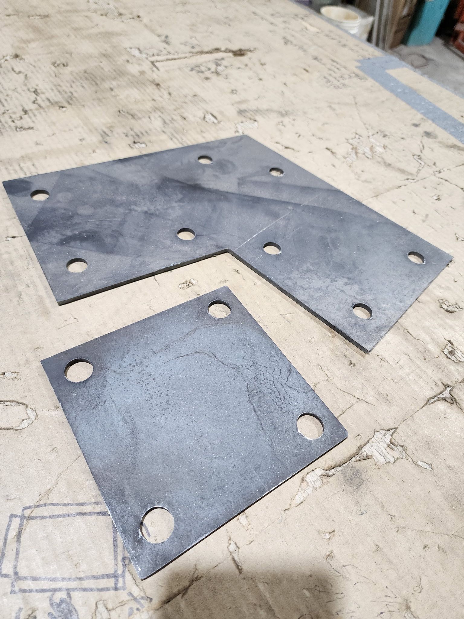 Two steel plates with drilled holes sit on a wooden surface. One is L-shaped; the other is square.