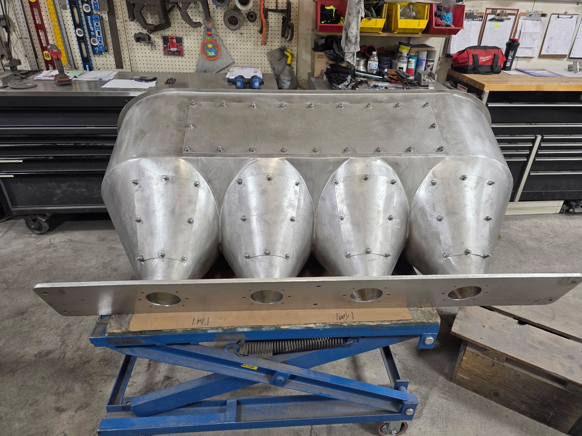 A silver intake manifold for an engine, on a lift table in a workshop.