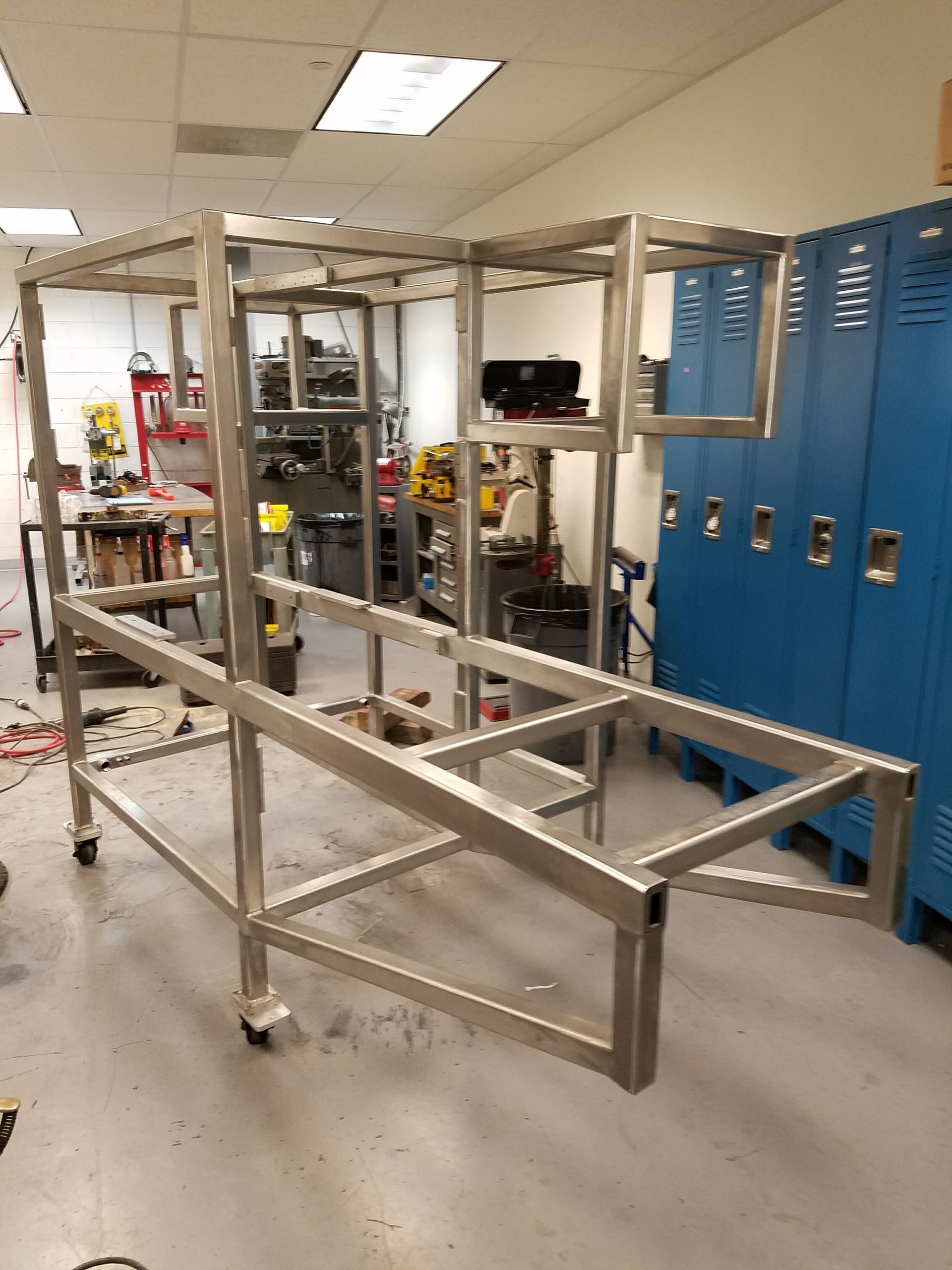 A large, silver metal frame on wheels, likely a cart or workstation, inside a workshop with blue lockers.