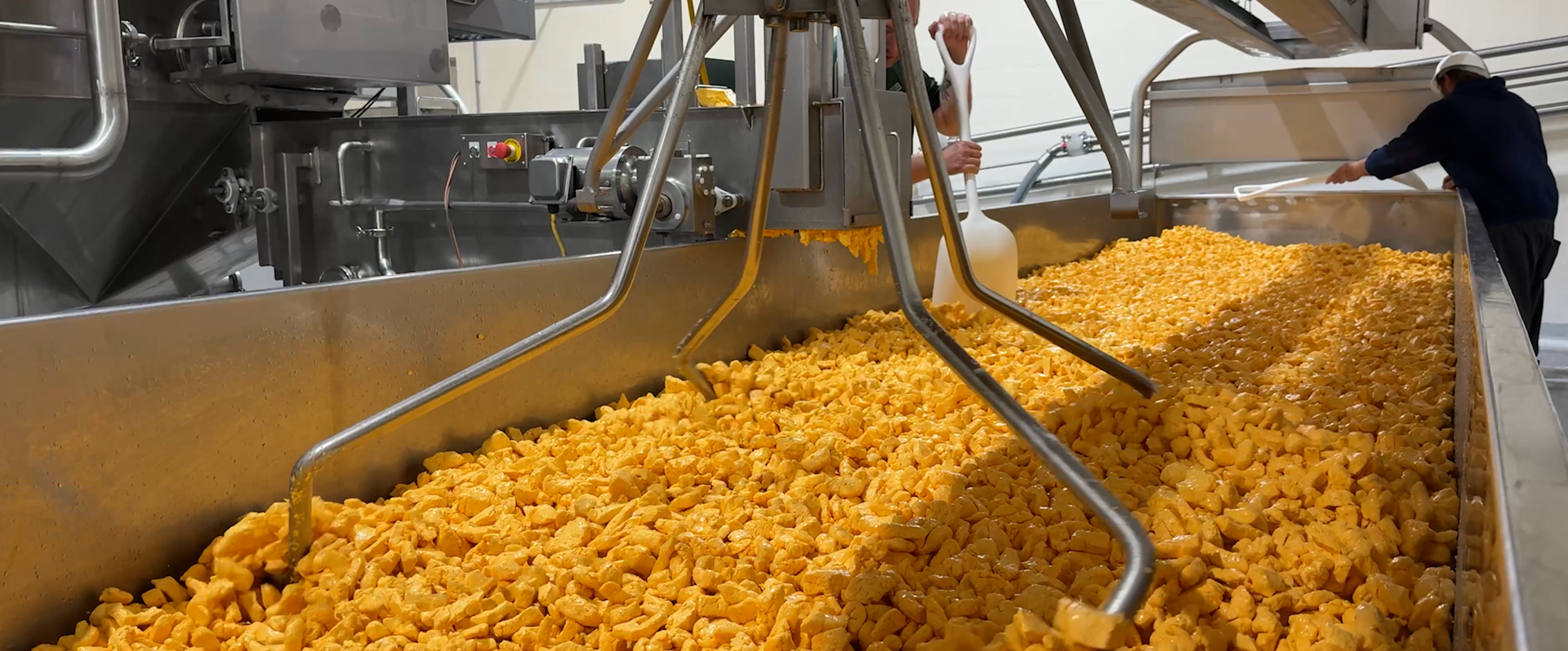 Large vat filled with orange food product, possibly cheese curds, with industrial machinery and a worker in the background.