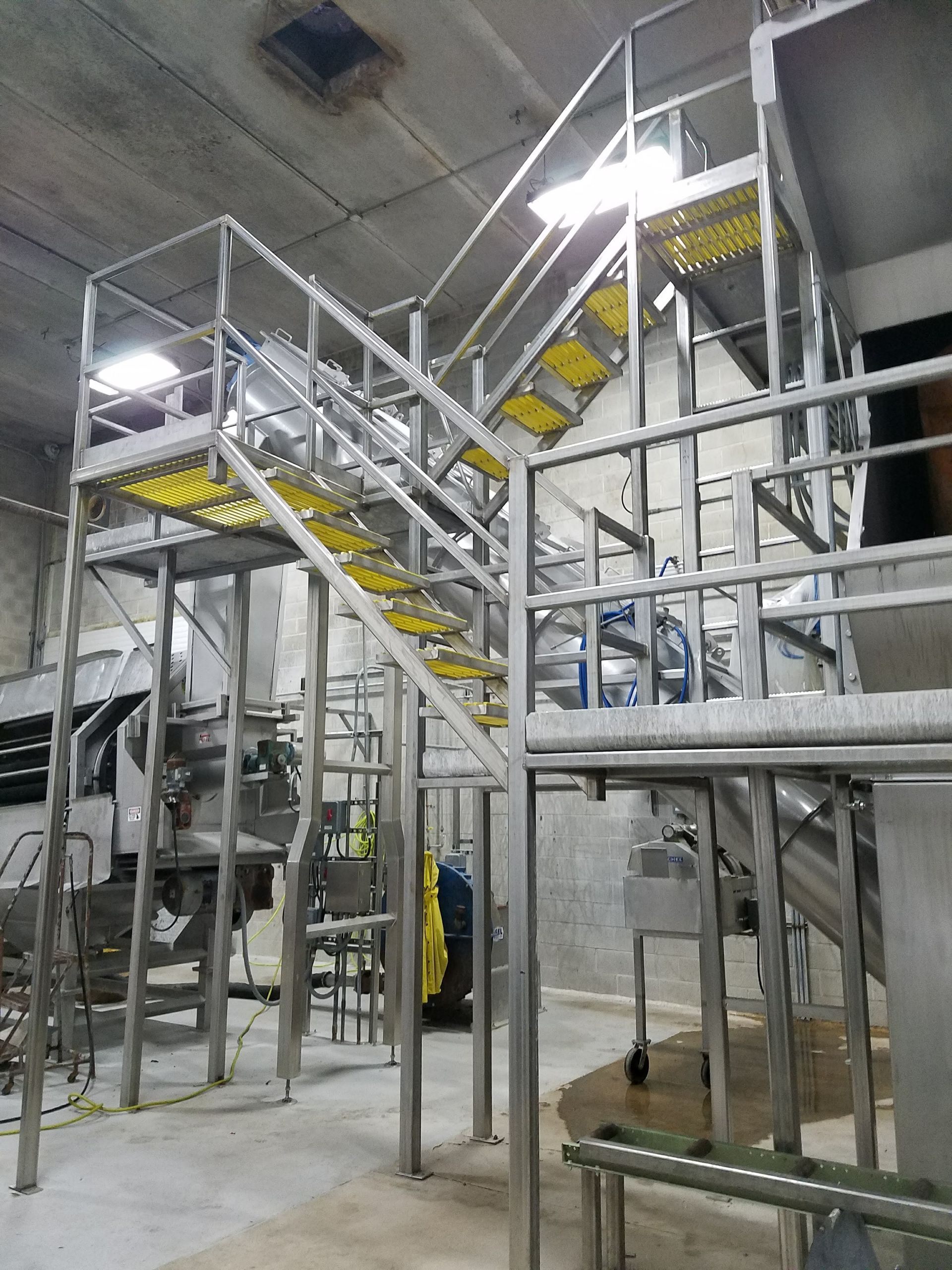 Stainless steel industrial staircase with yellow non-slip treads, inside a processing facility.
