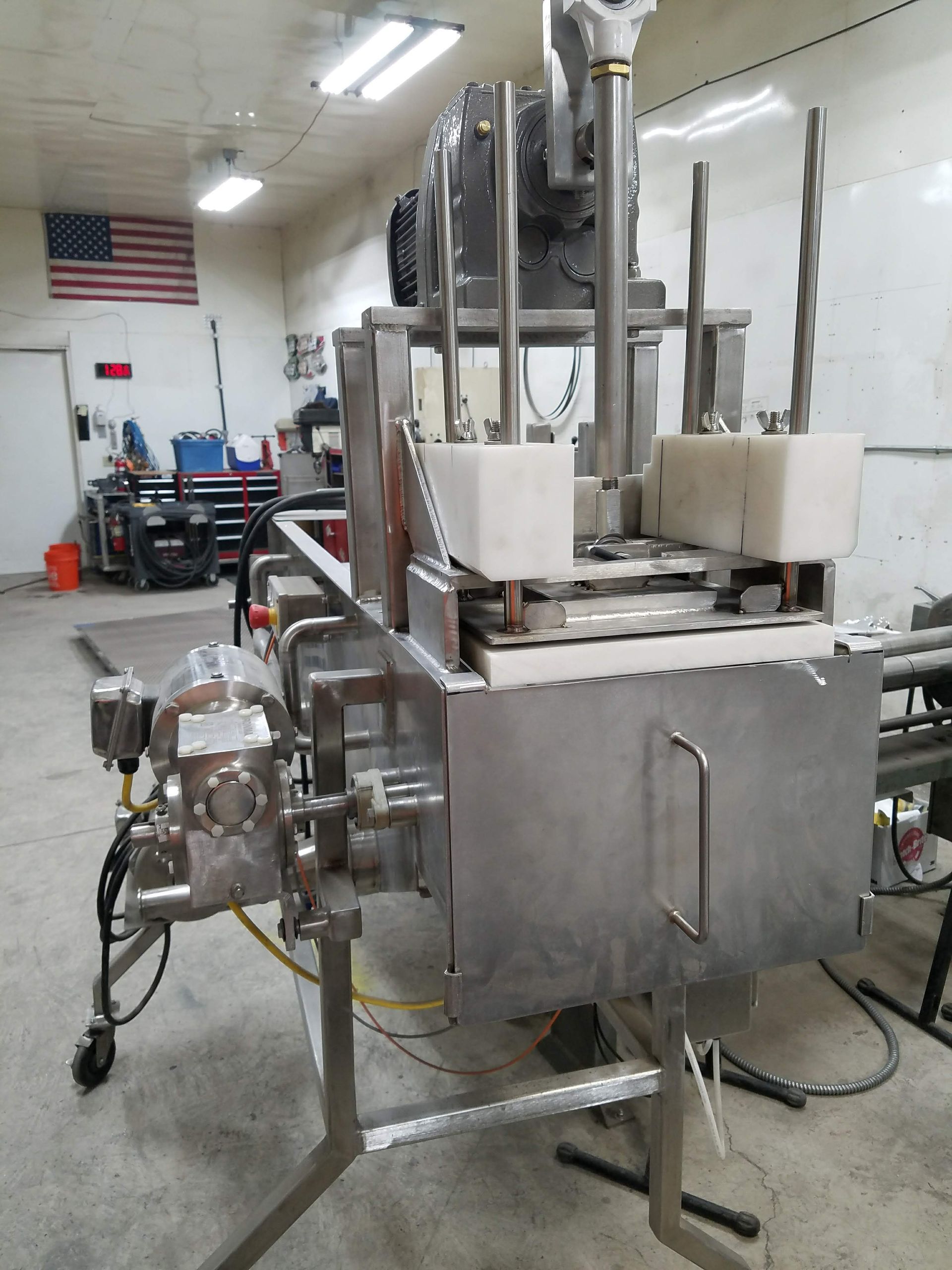 Stainless steel industrial machine in a workshop, possibly for food processing, with an American flag in the background.