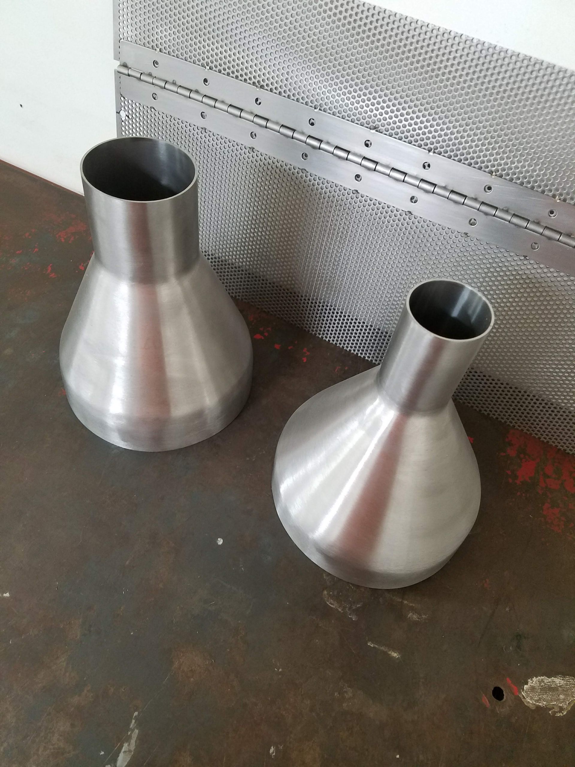Two stainless steel funnels on a rusty surface with a perforated metal sheet in the background.