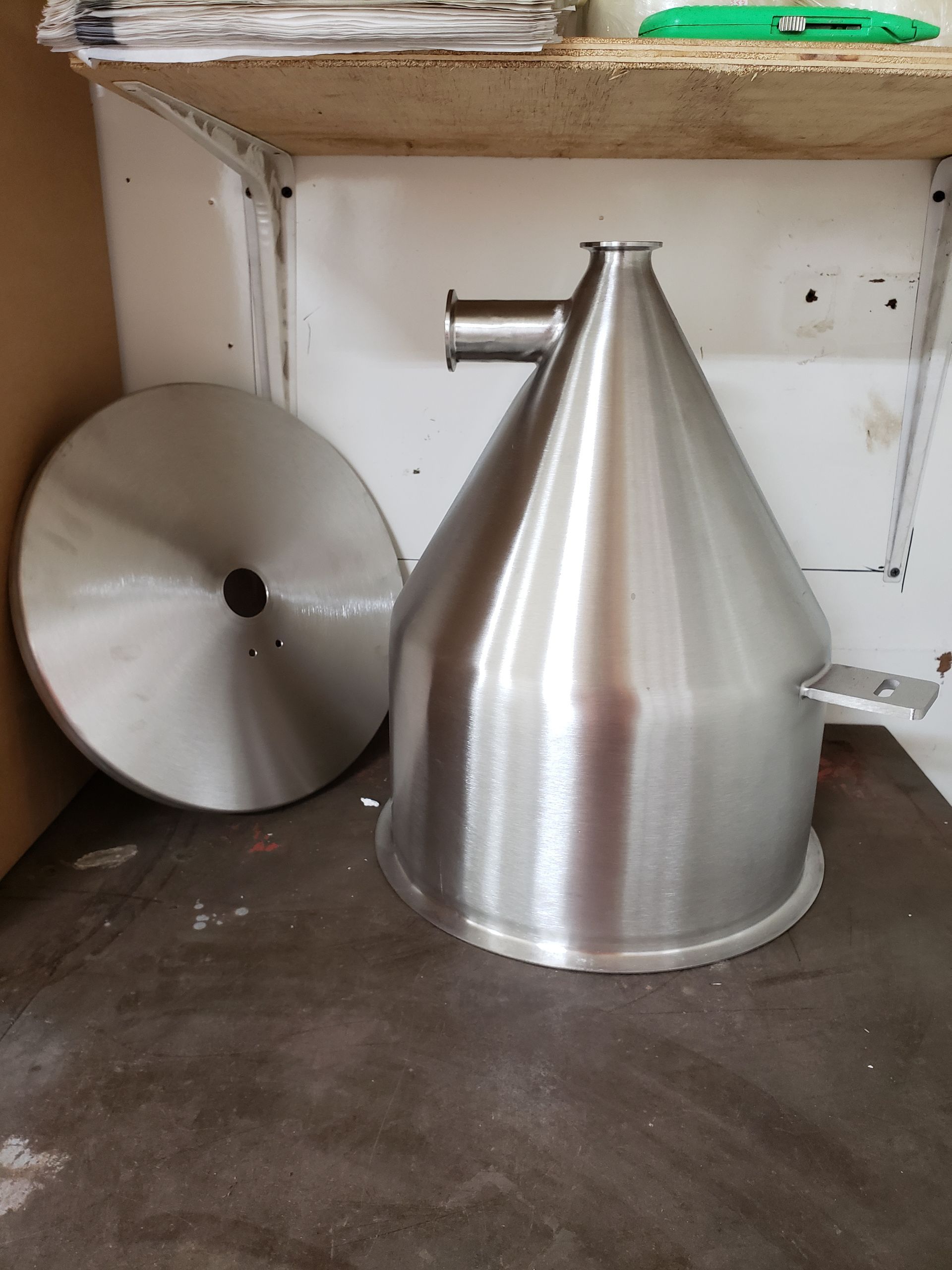 Stainless steel conical hopper with a lid, on a metal shelf.