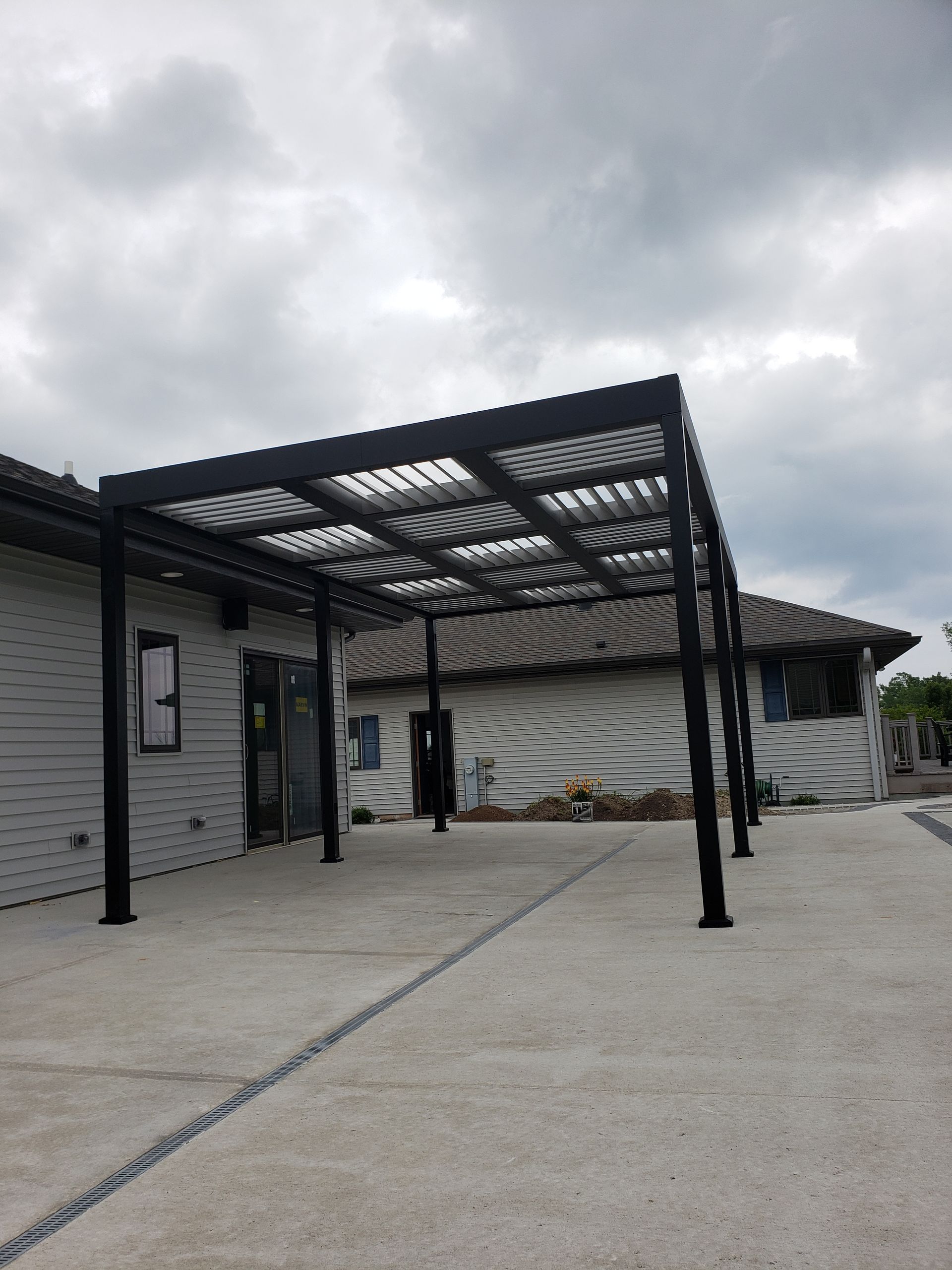 Black pergola attached to a white building, over a concrete patio, under a cloudy sky.