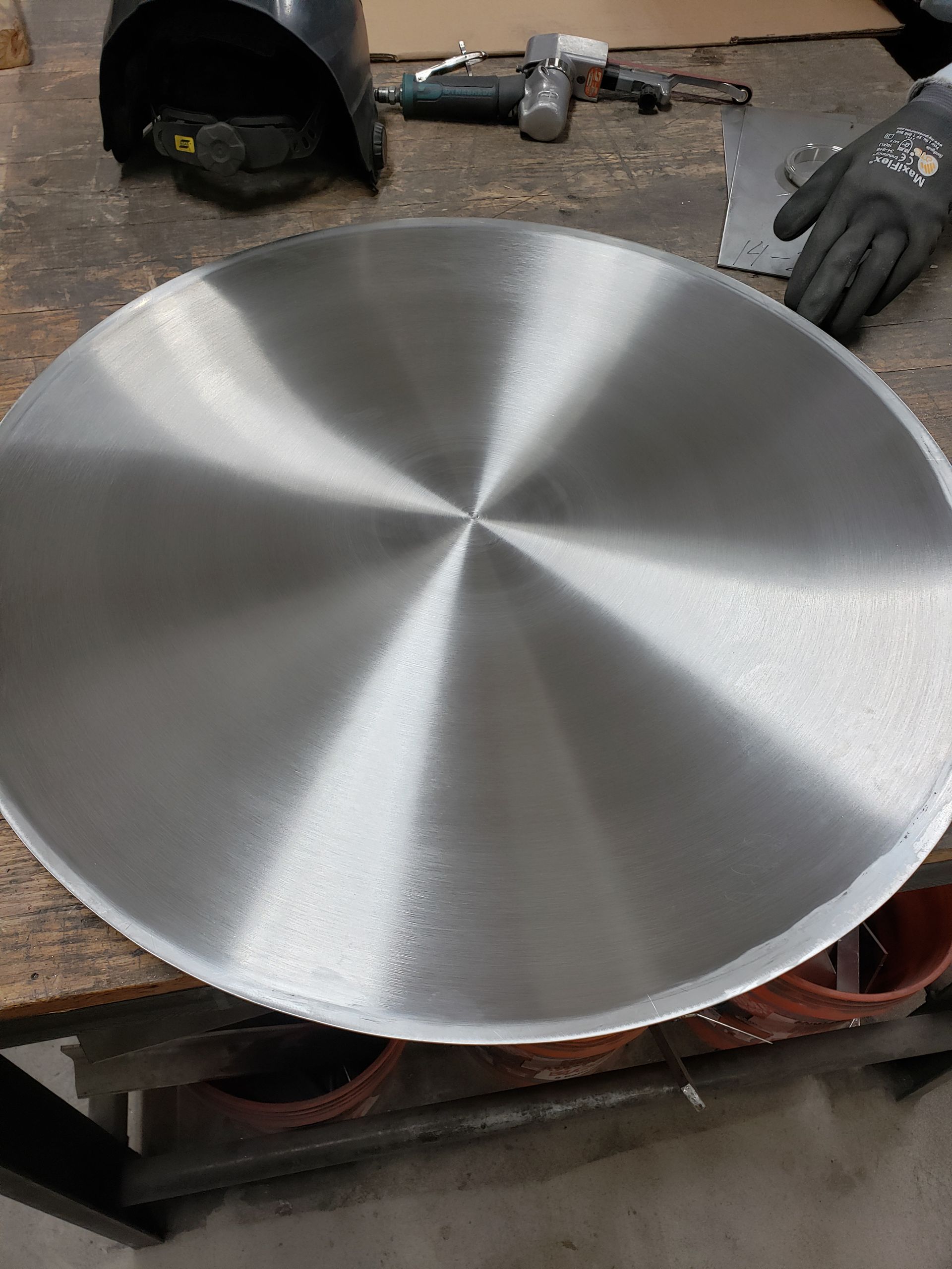 Circular, brushed-metal sheet on a work table with tools and a gloved hand in the background.