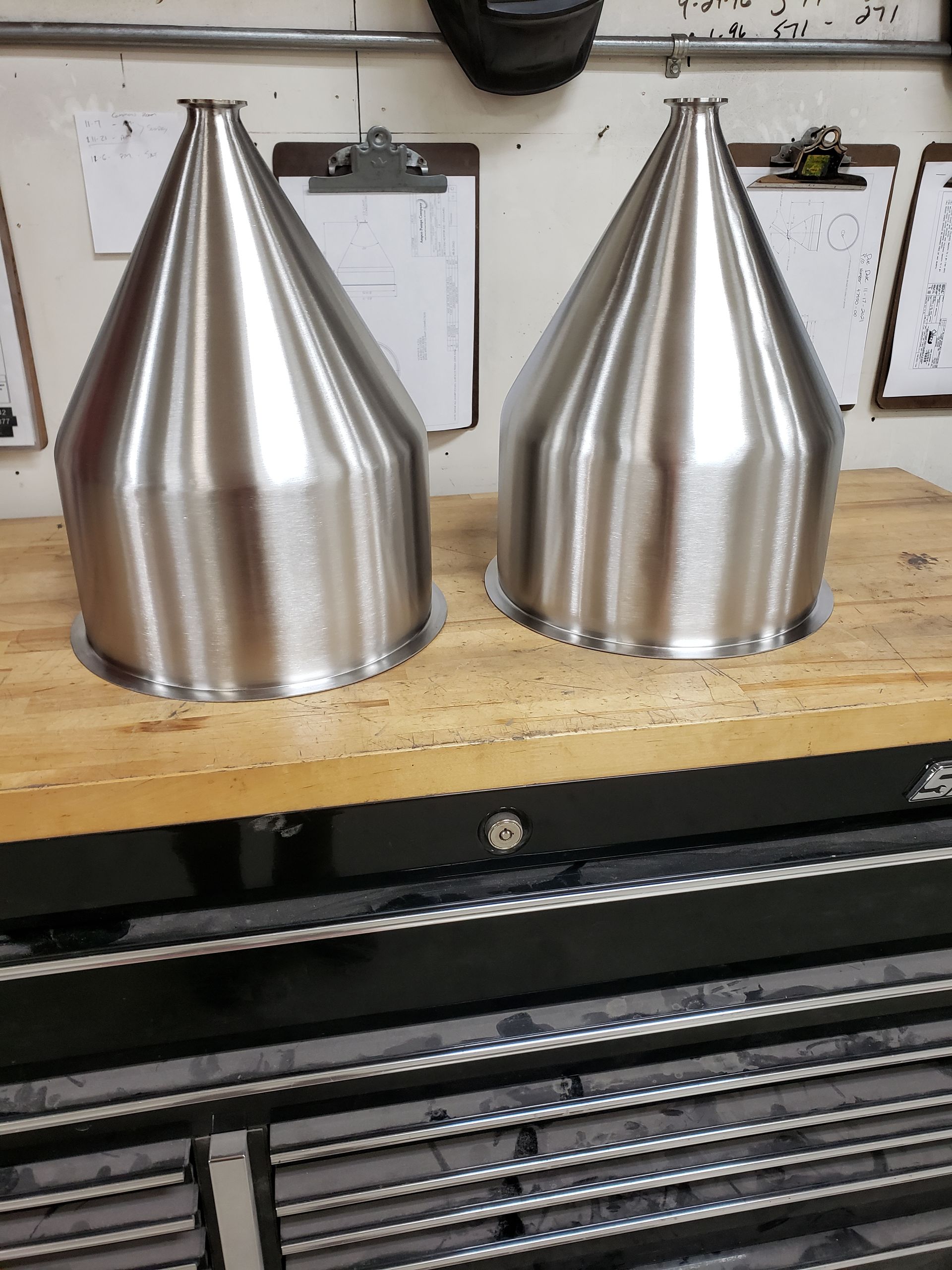 Two shiny, silver, cone-shaped metal objects sitting on a wooden work surface in a workshop.