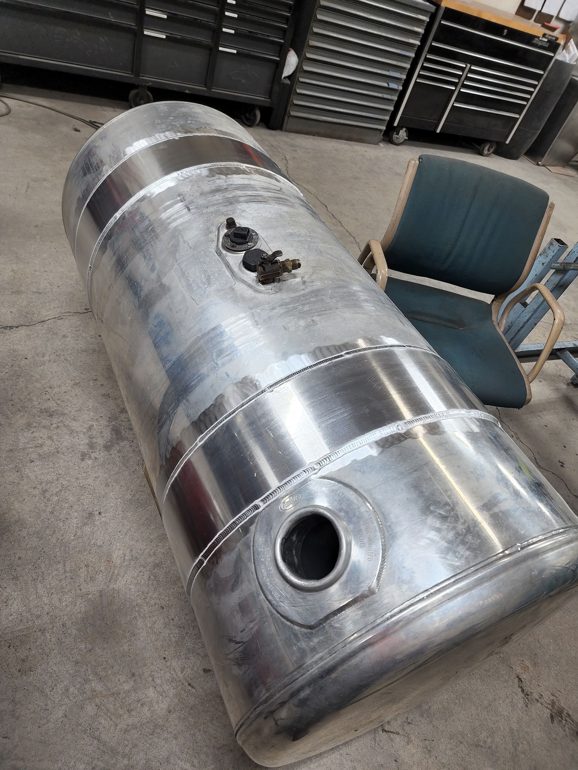 Silver metal cylindrical tank with fittings, on a concrete floor, near a chair.