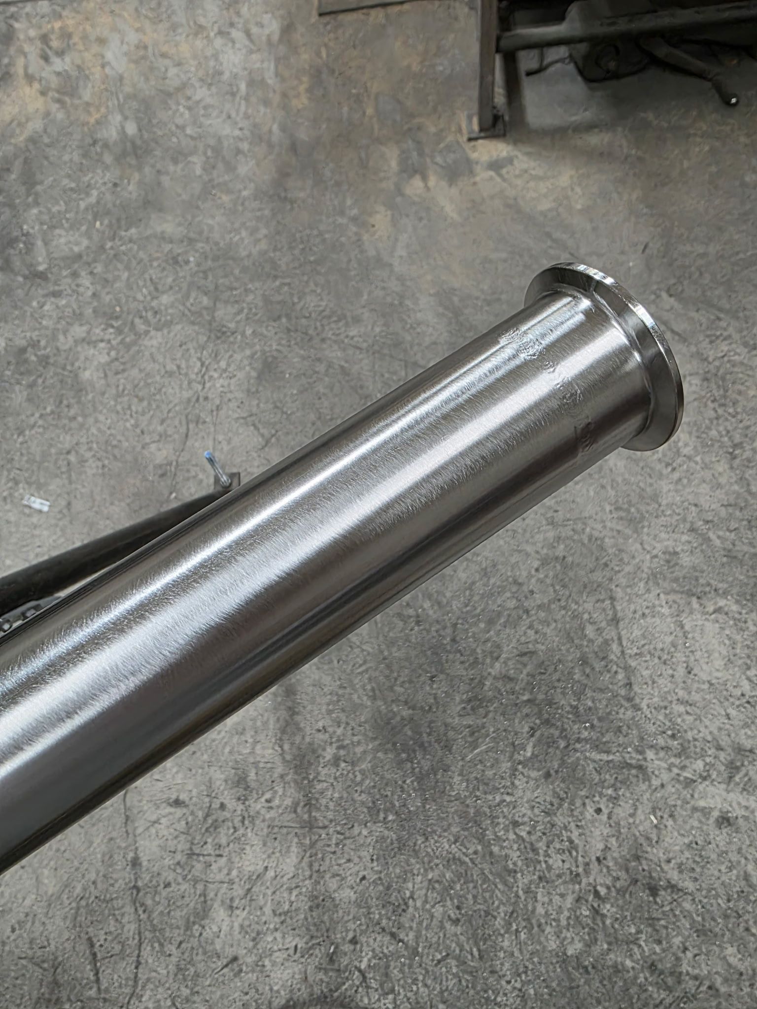Shiny metal pipe with a flange at one end, lying on a gray concrete surface.
