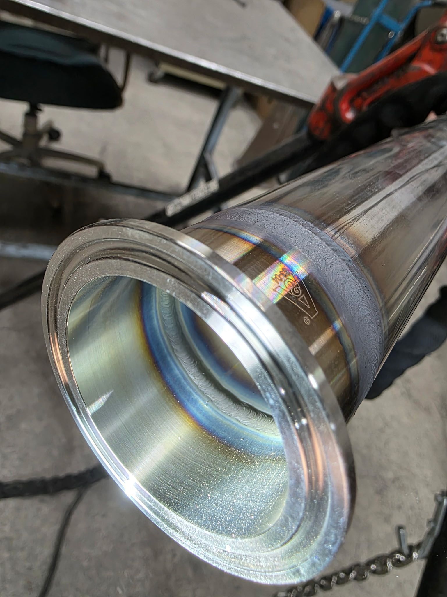 Close-up of welded metal tube with blue and yellow weld lines, held in a workshop setting