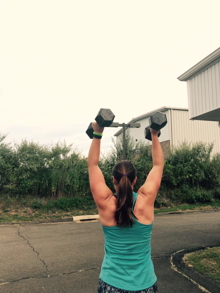 A woman is lifting dumbbells over her head in front of a building.
