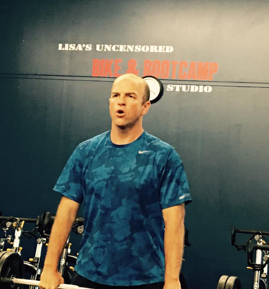 A man lifting a barbell in front of a sign that says lisa 's uncensored bike bootcamp studio