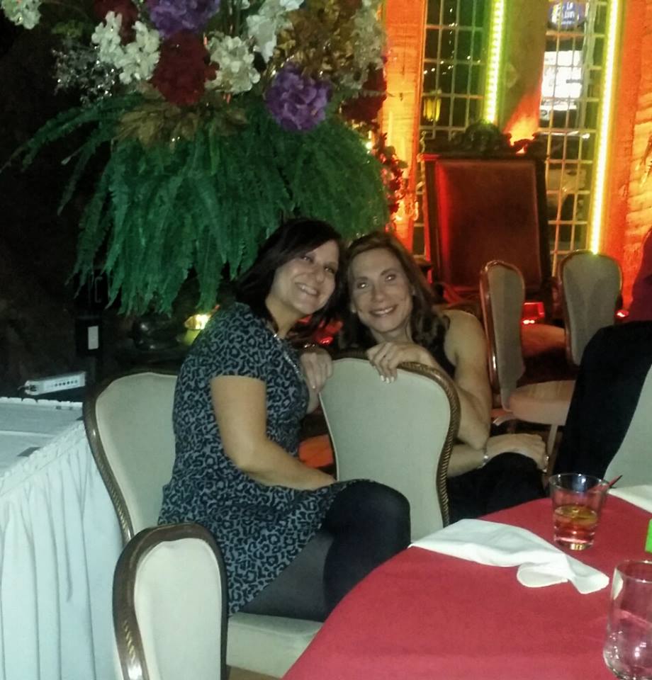Two women sit at a table with a vase of flowers in the background