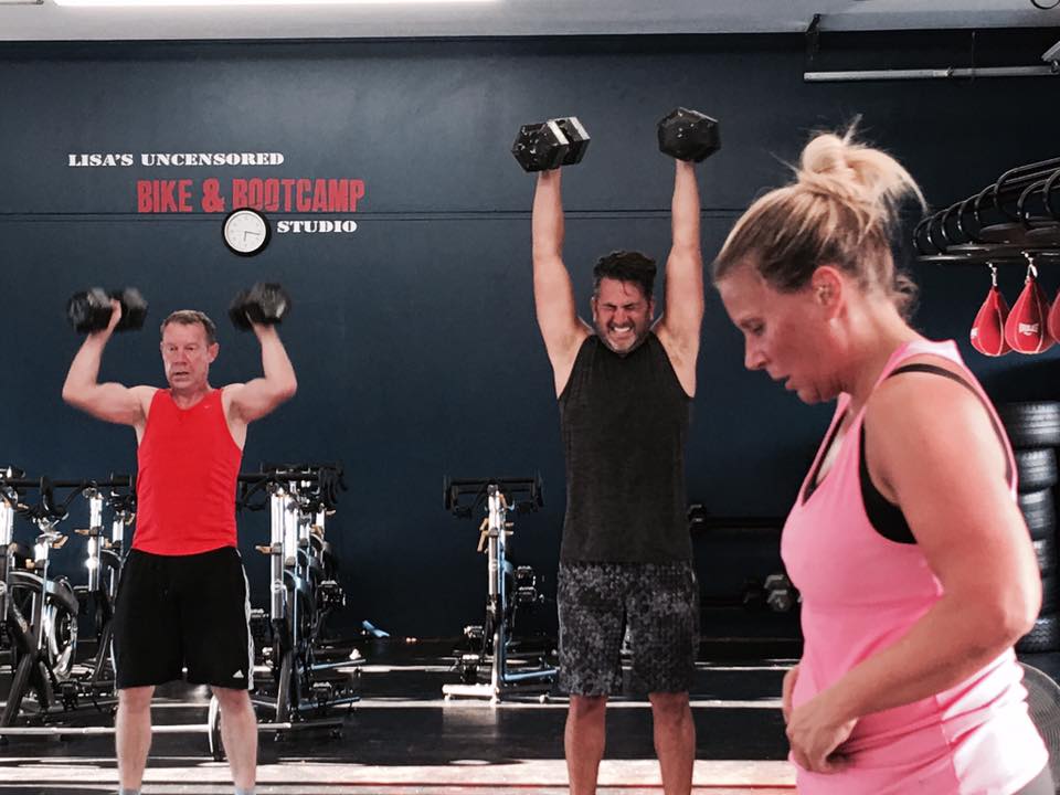 A man is lifting dumbbells in a gym while a woman looks on