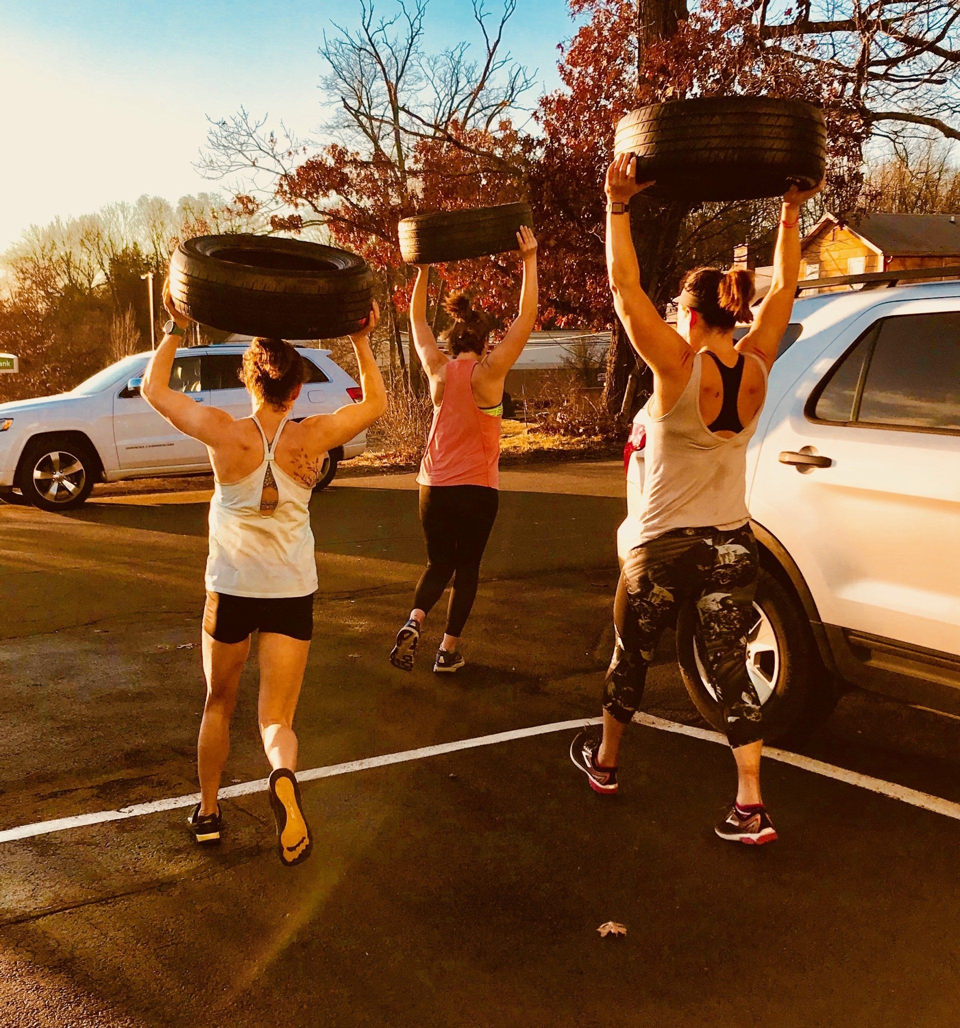 Three women are carrying tires over their heads in a parking lot