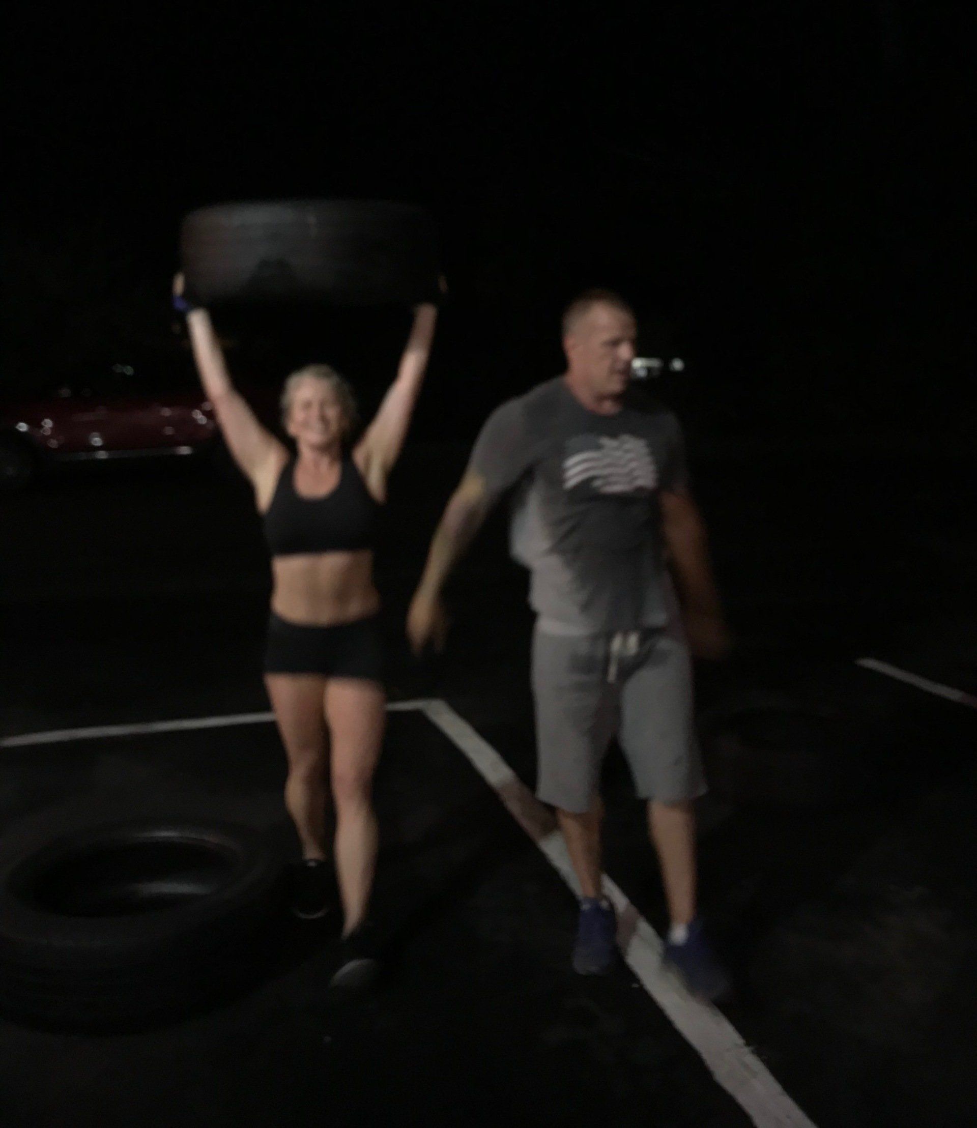 A man and a woman are lifting a tire in a parking lot
