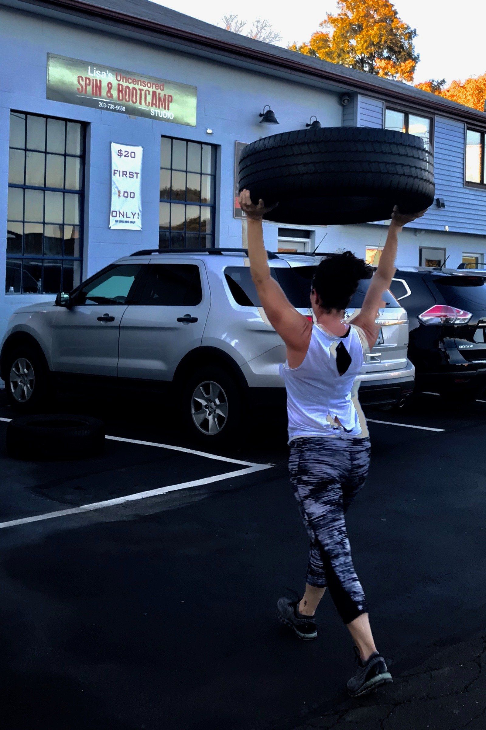 A woman is carrying a tire over her head in a parking lot.