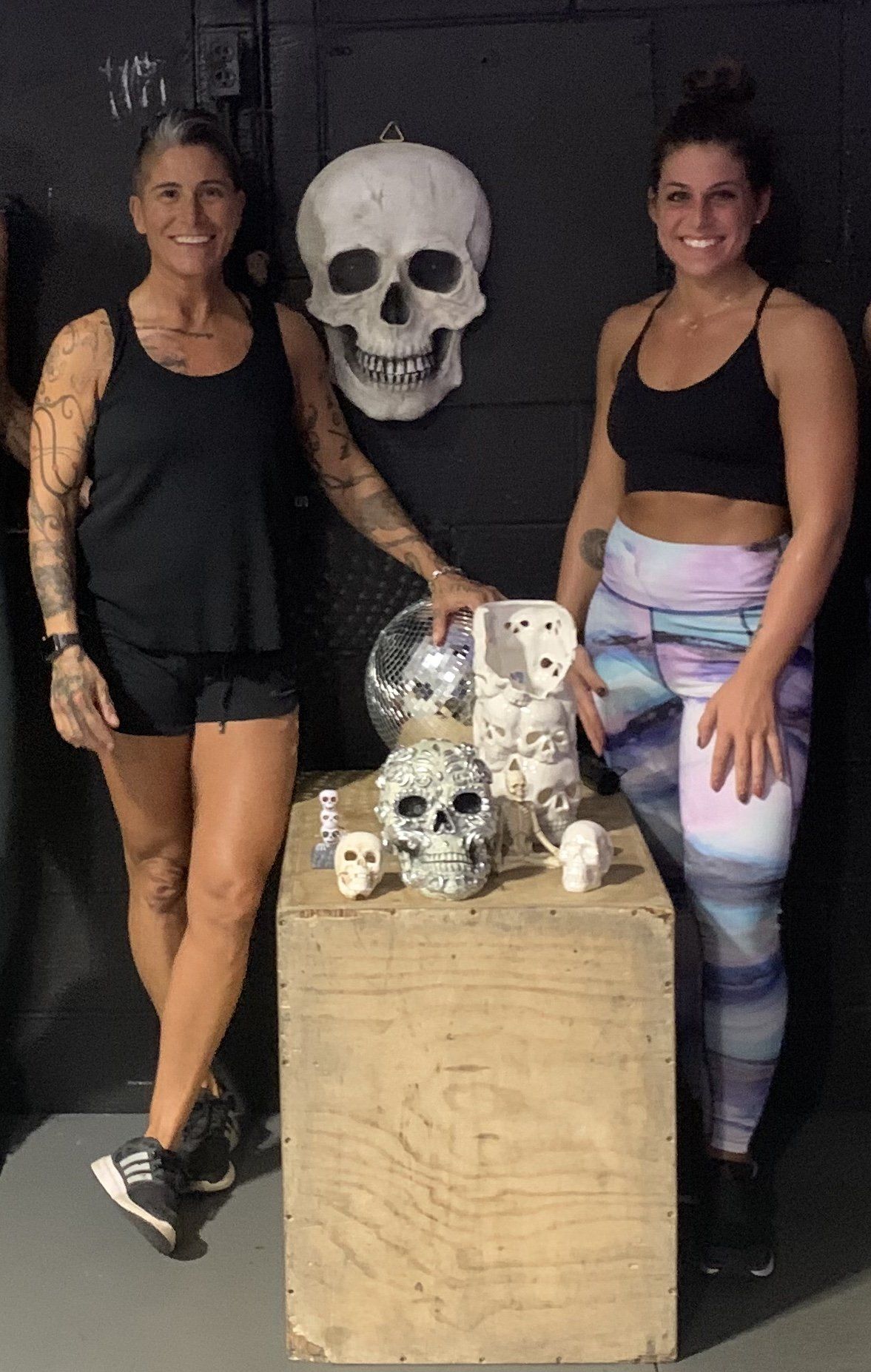 Two women are standing next to a wooden box with skulls on it.