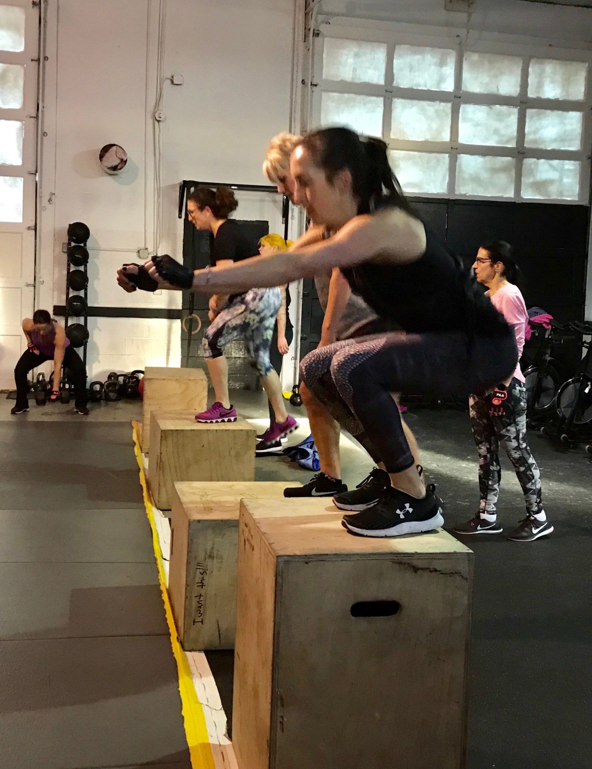 A woman is squatting on a wooden box in a gym.