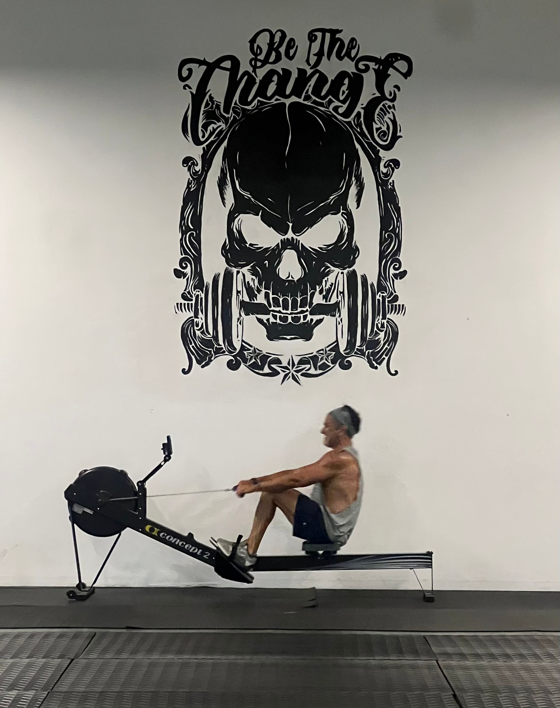 A man is sitting on a rowing machine in front of a wall that says be the change.