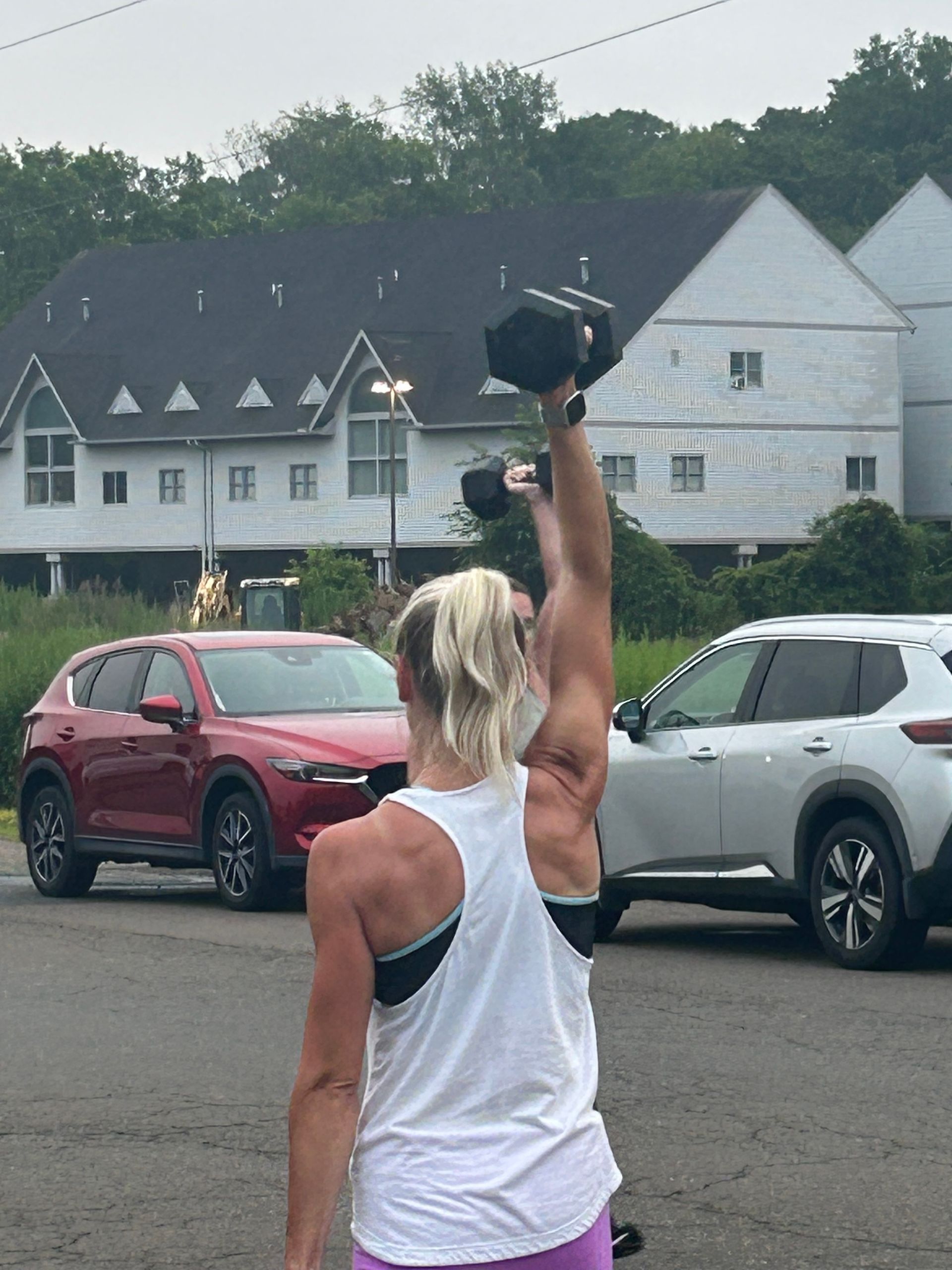 Person in white tank top raises a black dumbbell in a parking lot with cars and houses behind