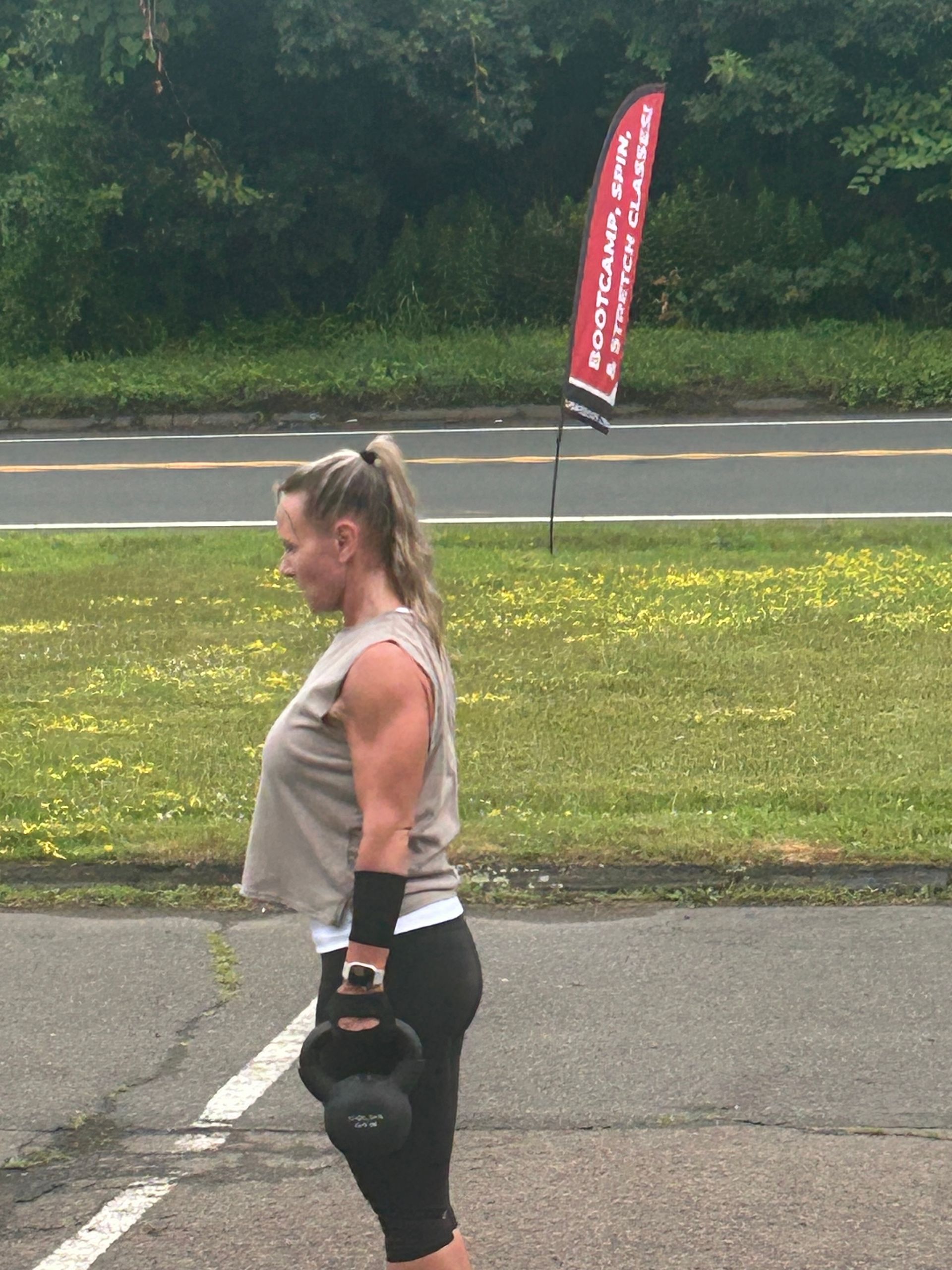Woman in athletic gear holding a kettlebell outdoors on a road near a grassy field