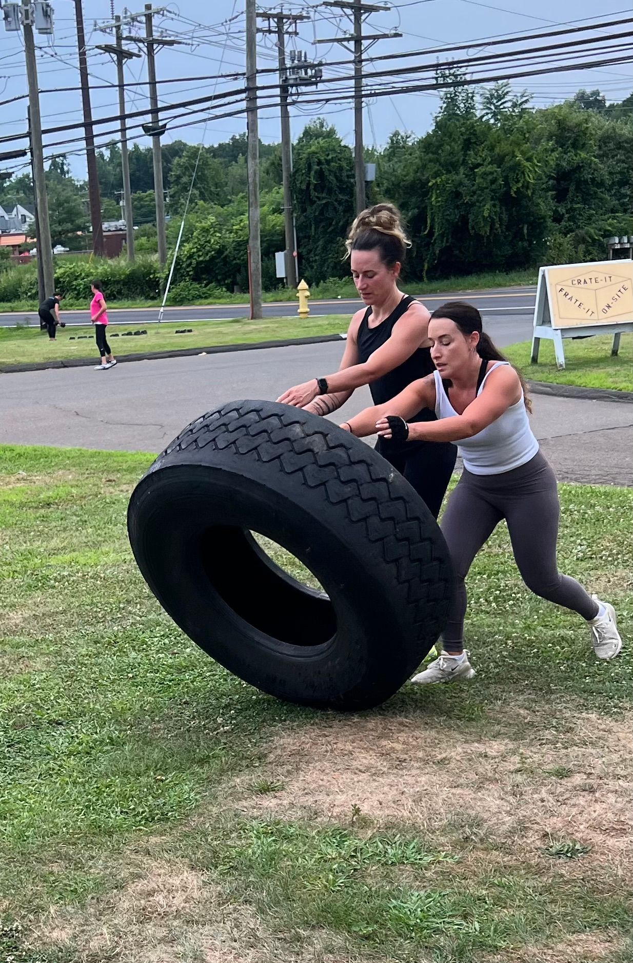Two women are pushing a large tire in a field.