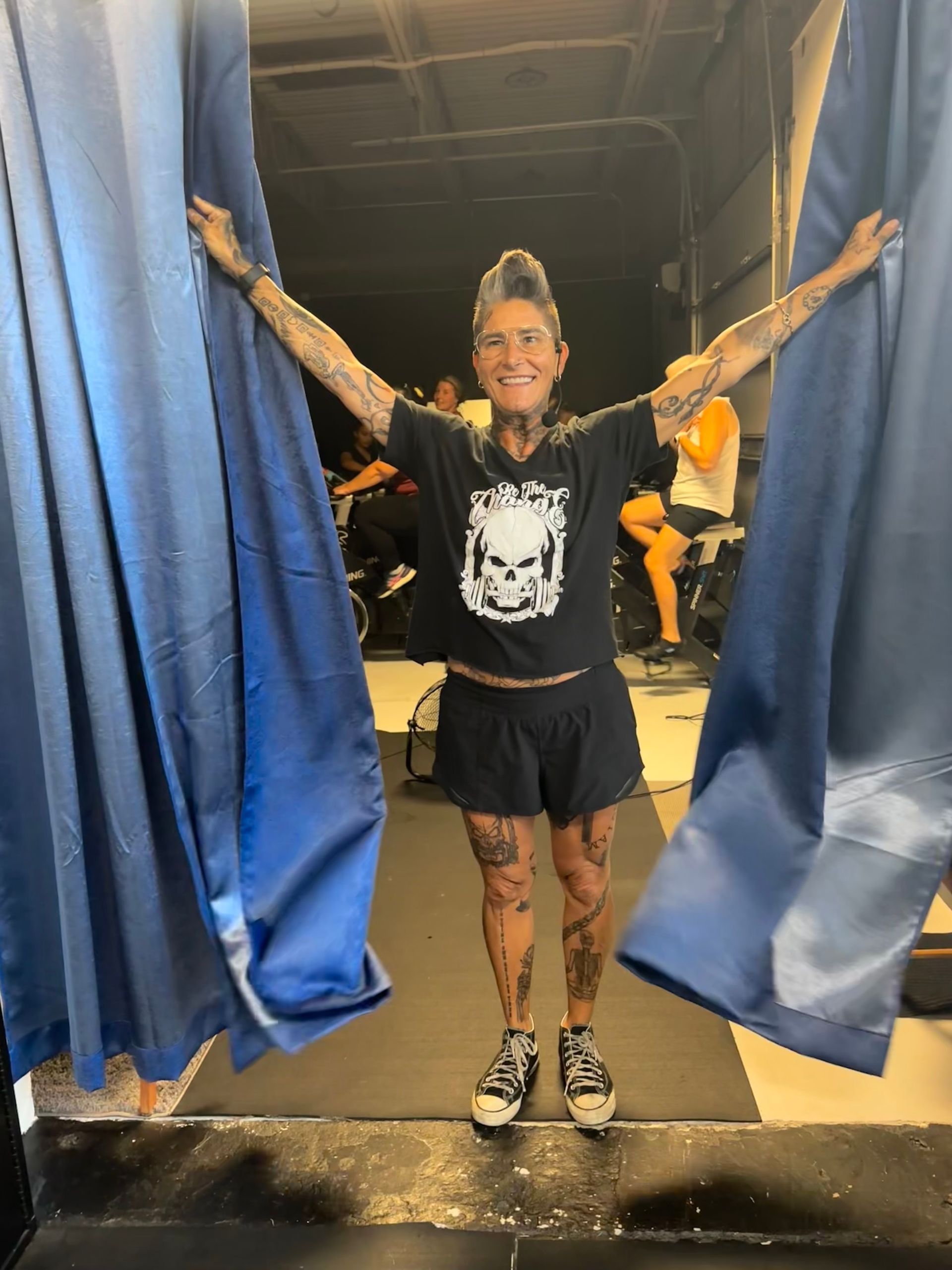 A woman in a black shirt with a skull on it is standing in front of a blue curtain.