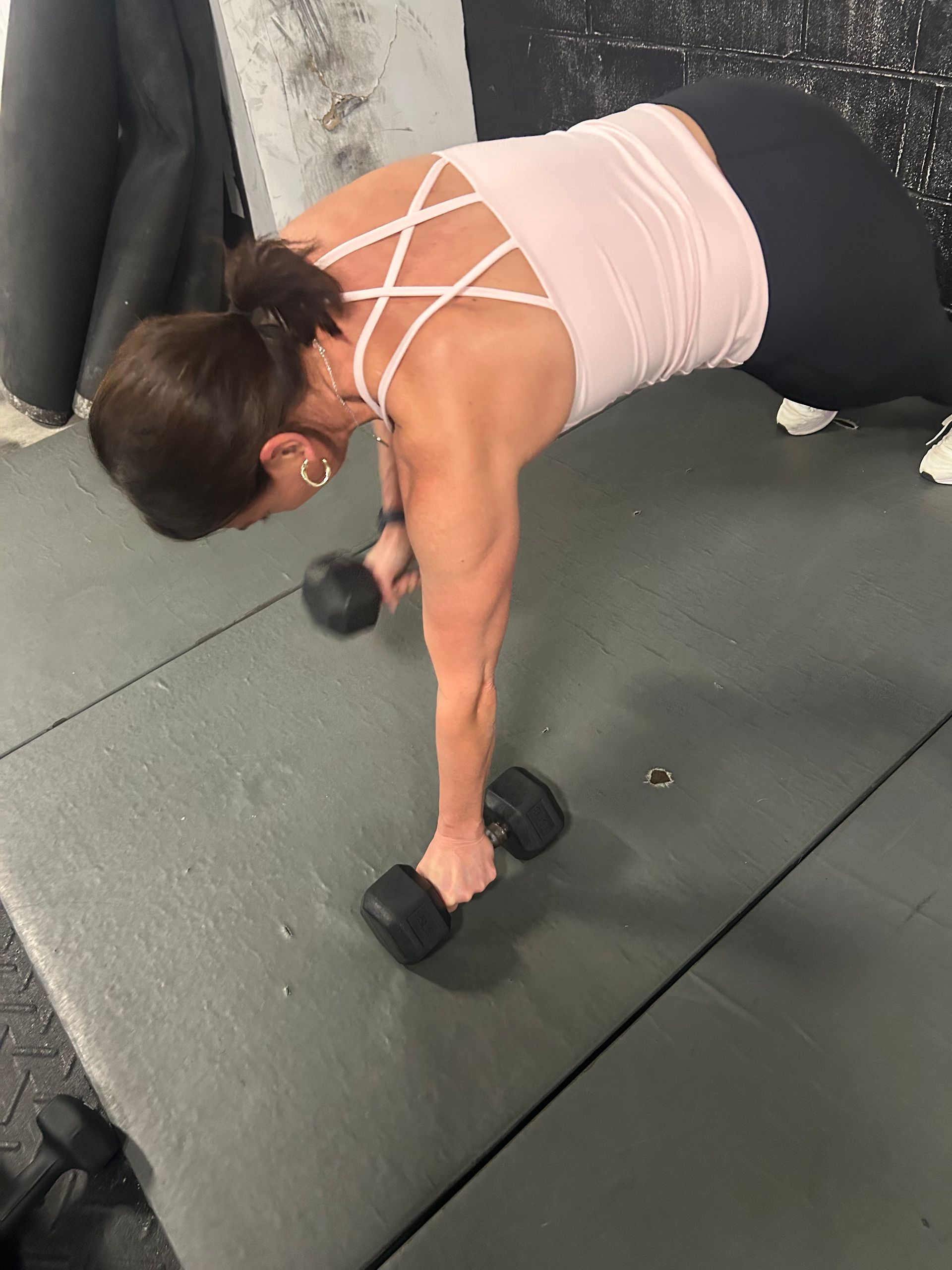 Person doing a dumbbell plank row on a gym floor, wearing a pink top and black leggings