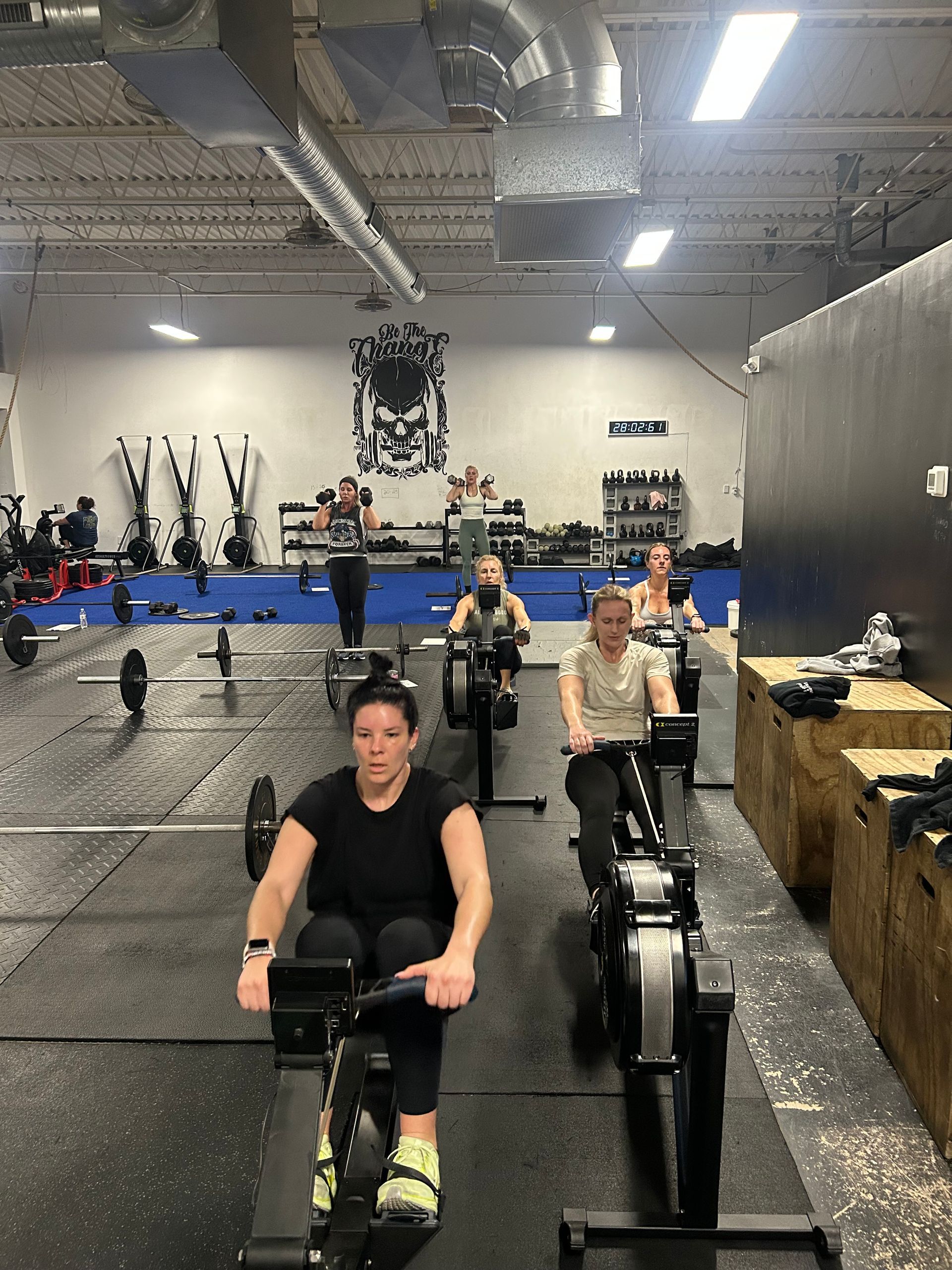 People rowing on machines in a large gym with black walls and overhead industrial lighting