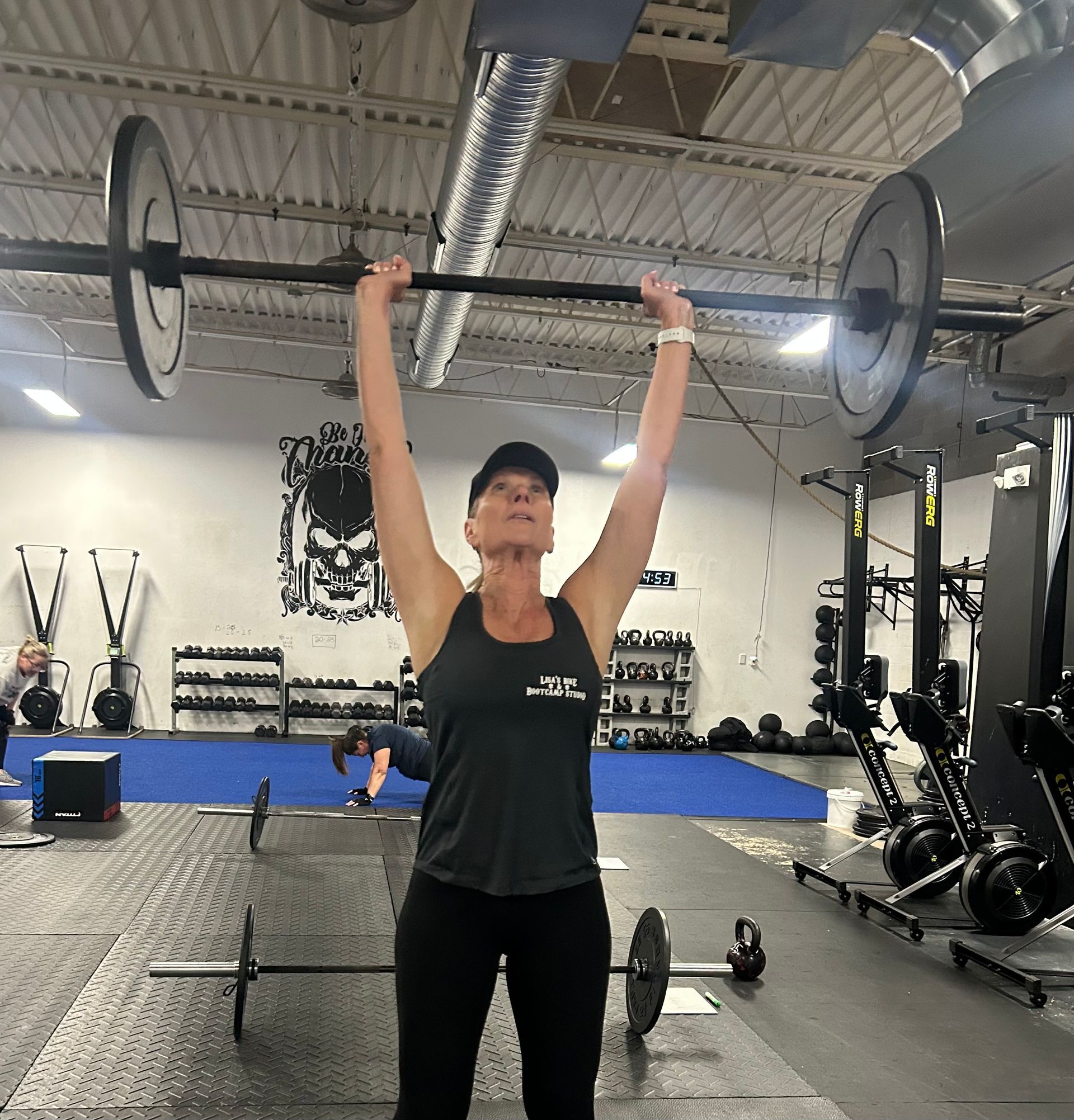 Person lifting a barbell overhead in a gym with black flooring and industrial ceiling ducts