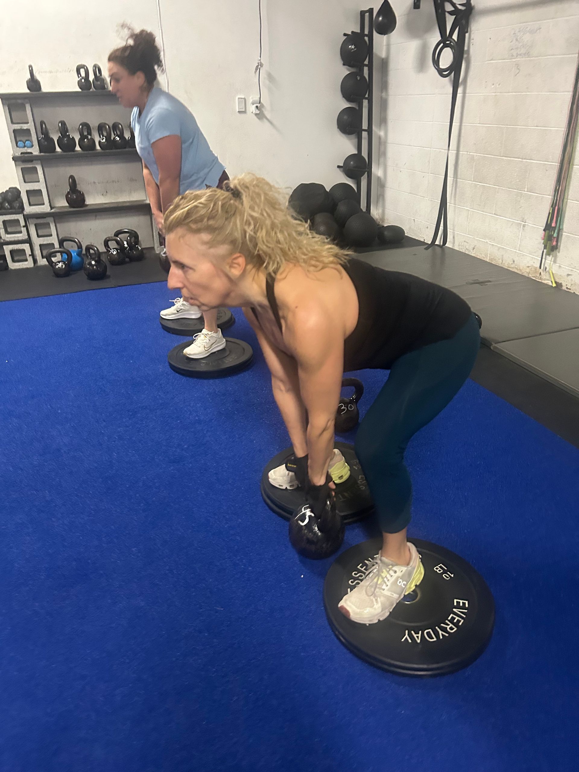 Two people doing kettlebell deadlifts in a gym on blue flooring.