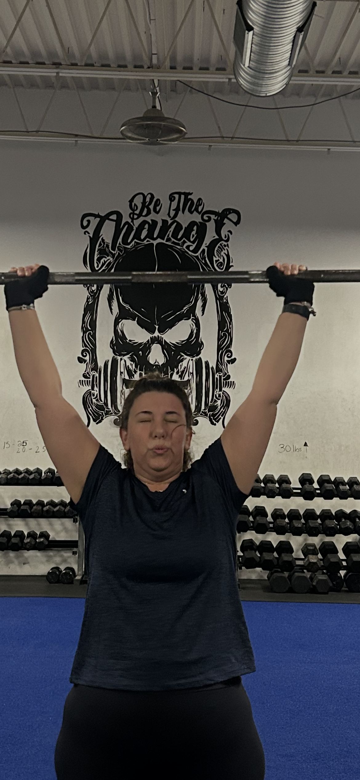 Person lifting a barbell overhead in a gym, standing on a blue floor with weights behind them