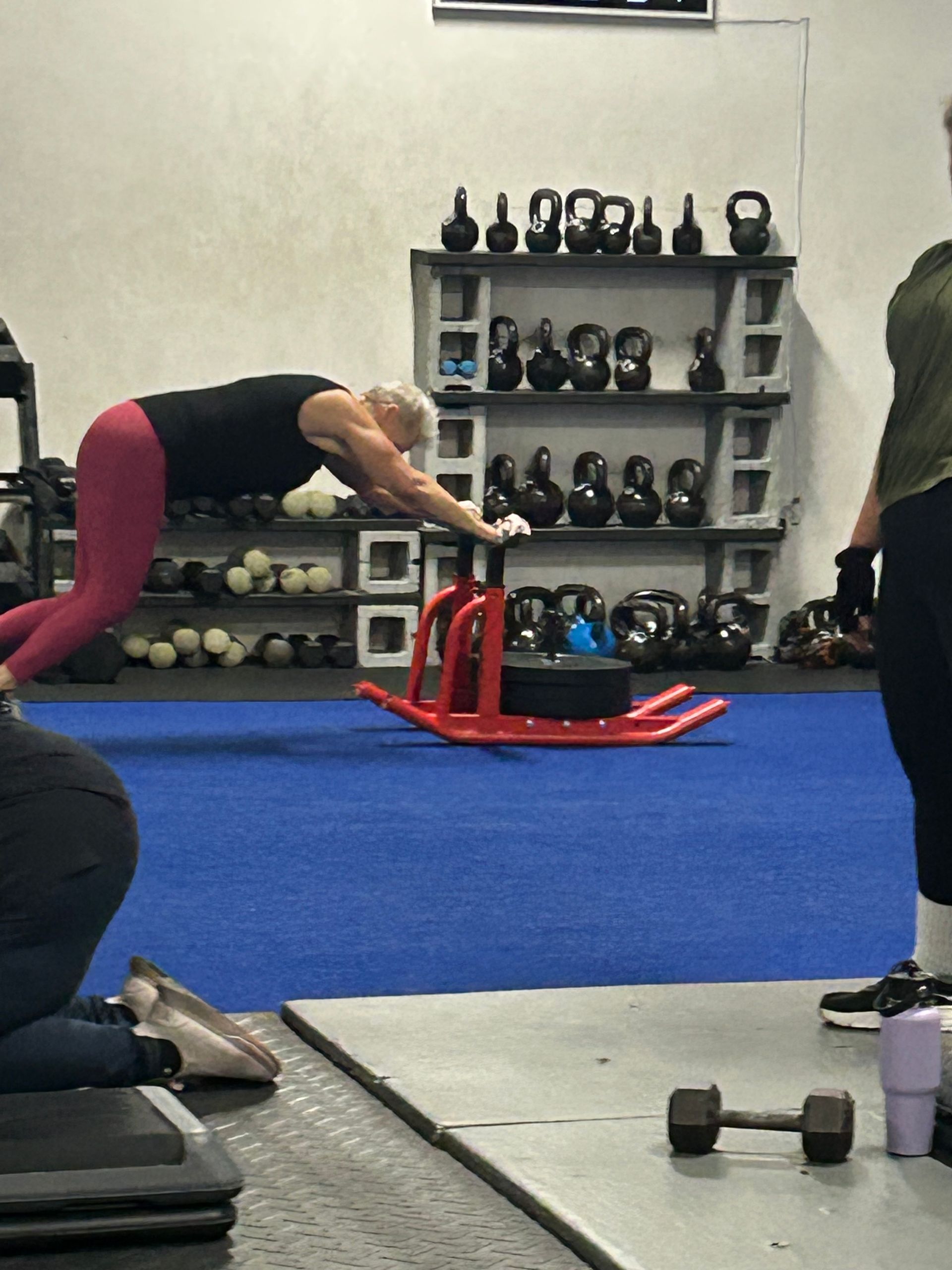Gym scene with a person leaning over a red sled on a blue floor, kettlebells on shelves in the background.