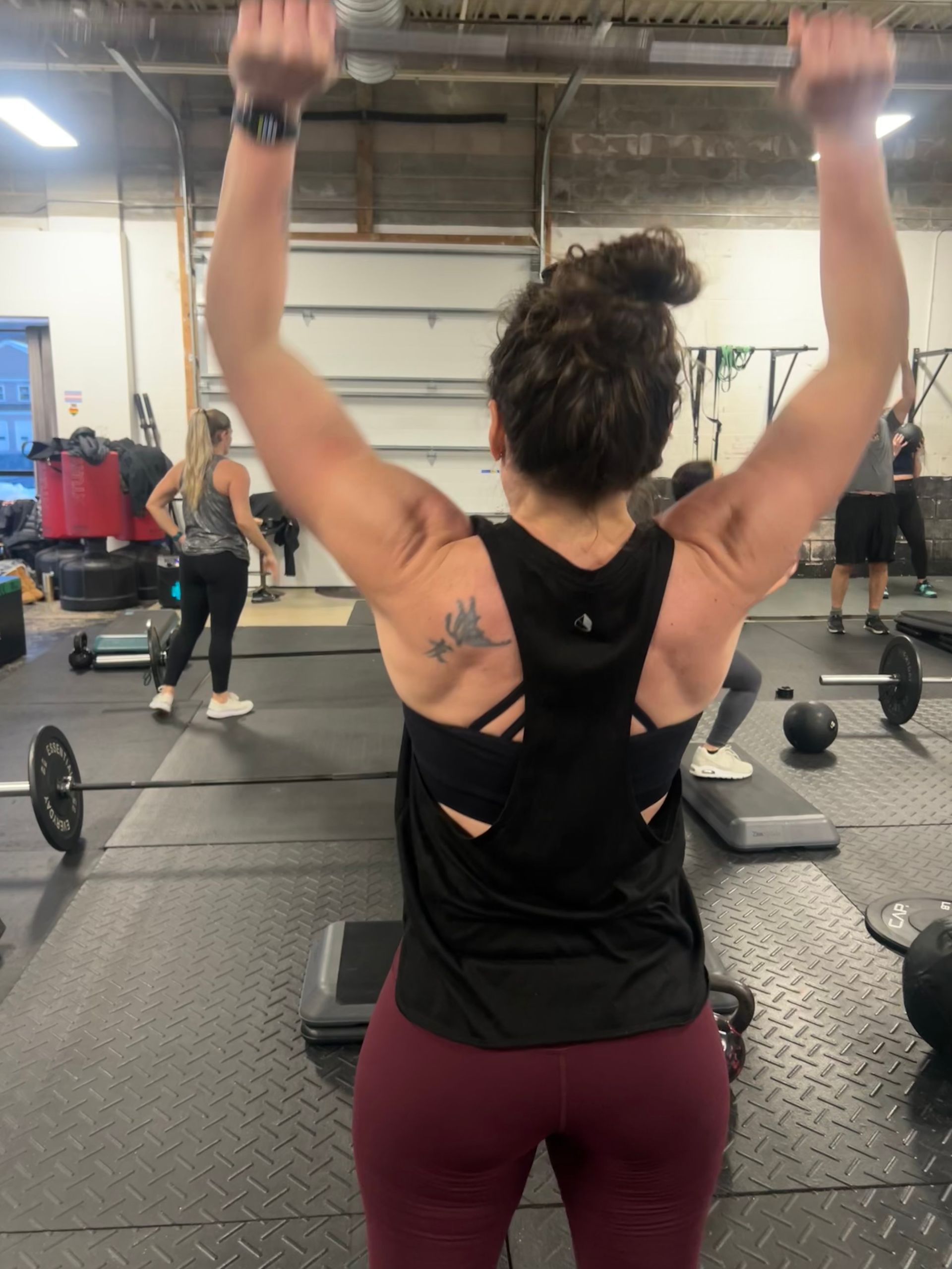 Woman lifting dumbbells overhead in a gym, seen from behind, wearing a black tank top and maroon leggings.