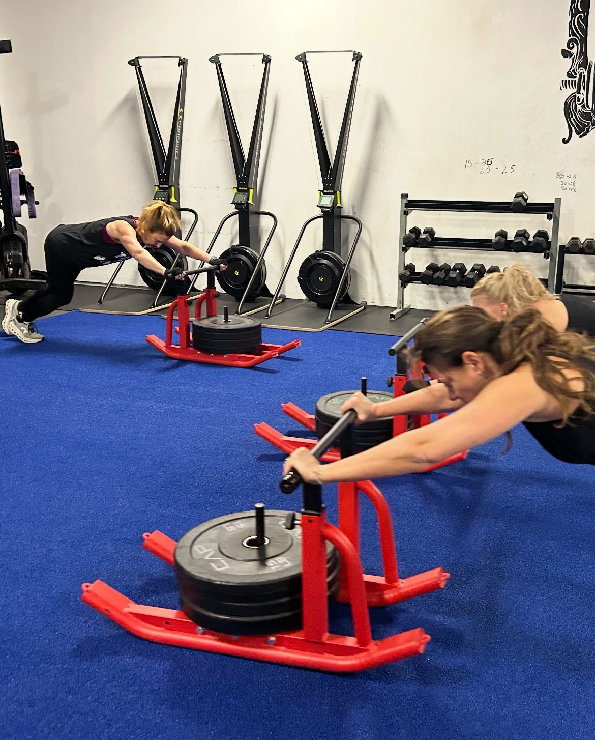 People pushing red sleds in a gym with blue turf and rowing machines in the background