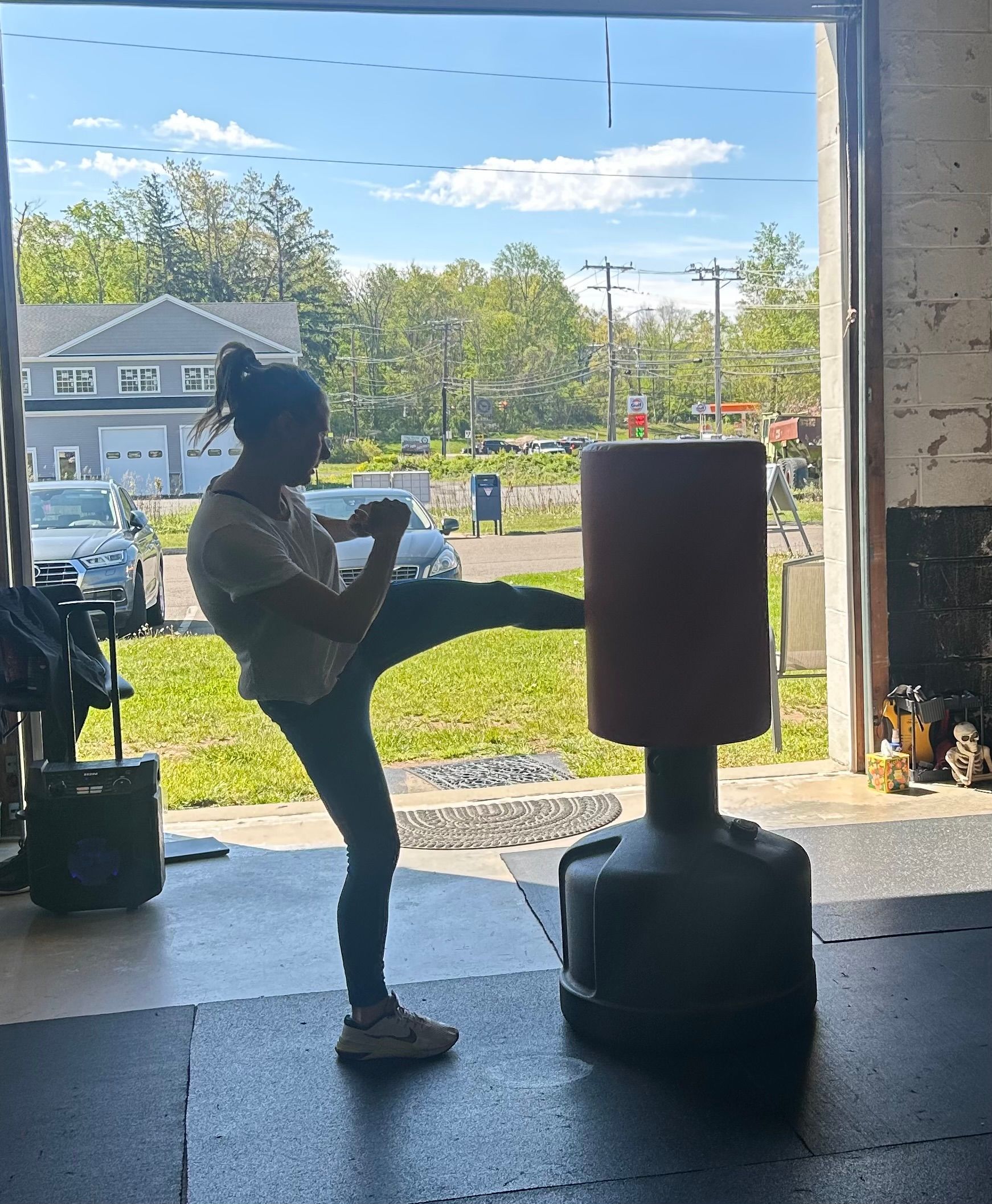 Person doing a front kick at a punching bag in a gym doorway, with daylight outside.