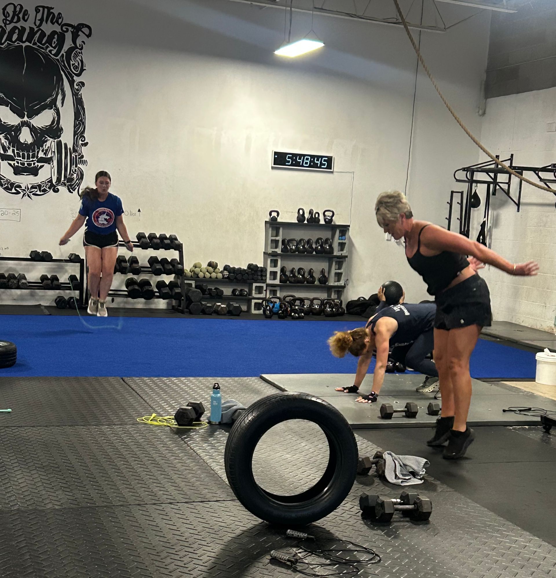 Gym with people doing strength exercises: kettlebell, tire, and dumbbells on the floor.
