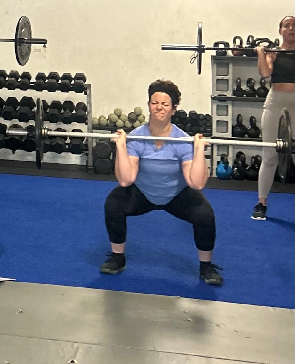 A person squats with a barbell in a gym with dumbbells and weights in the background.