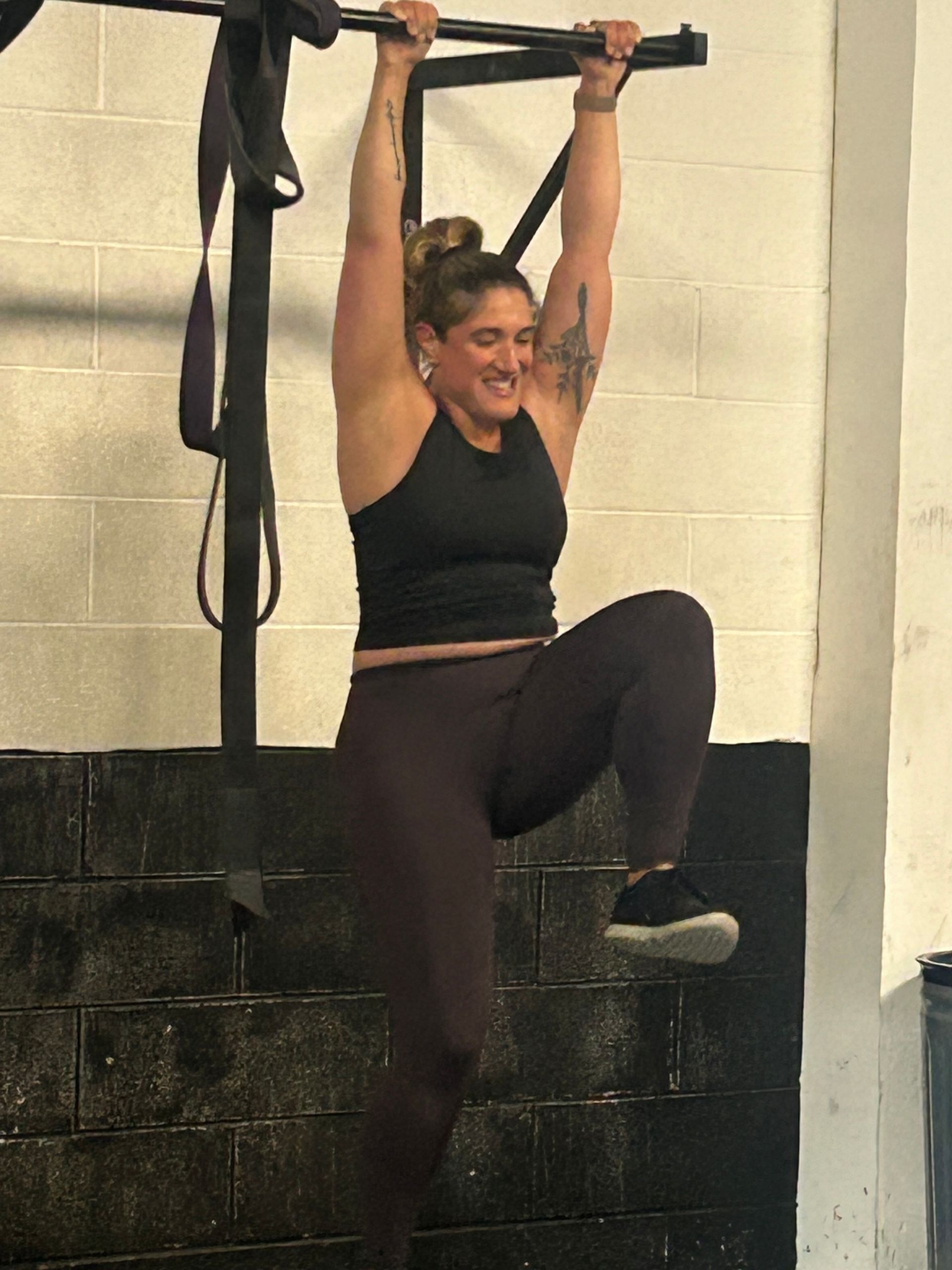 Woman hanging from a pull-up bar in a gym, one knee raised, smiling.