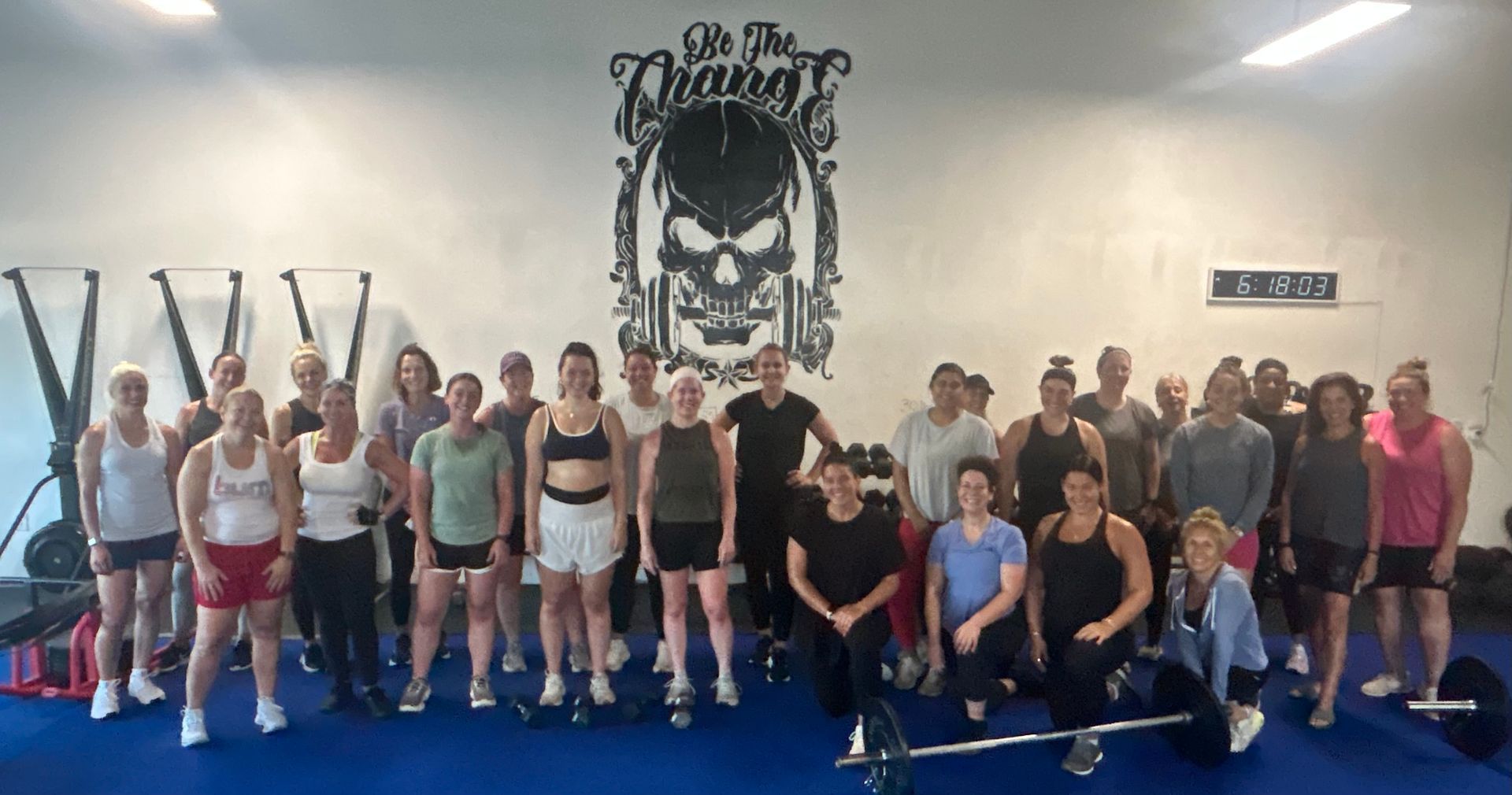 Group fitness class posing in a gym, with a skull logo on the wall and blue mats on the floor.