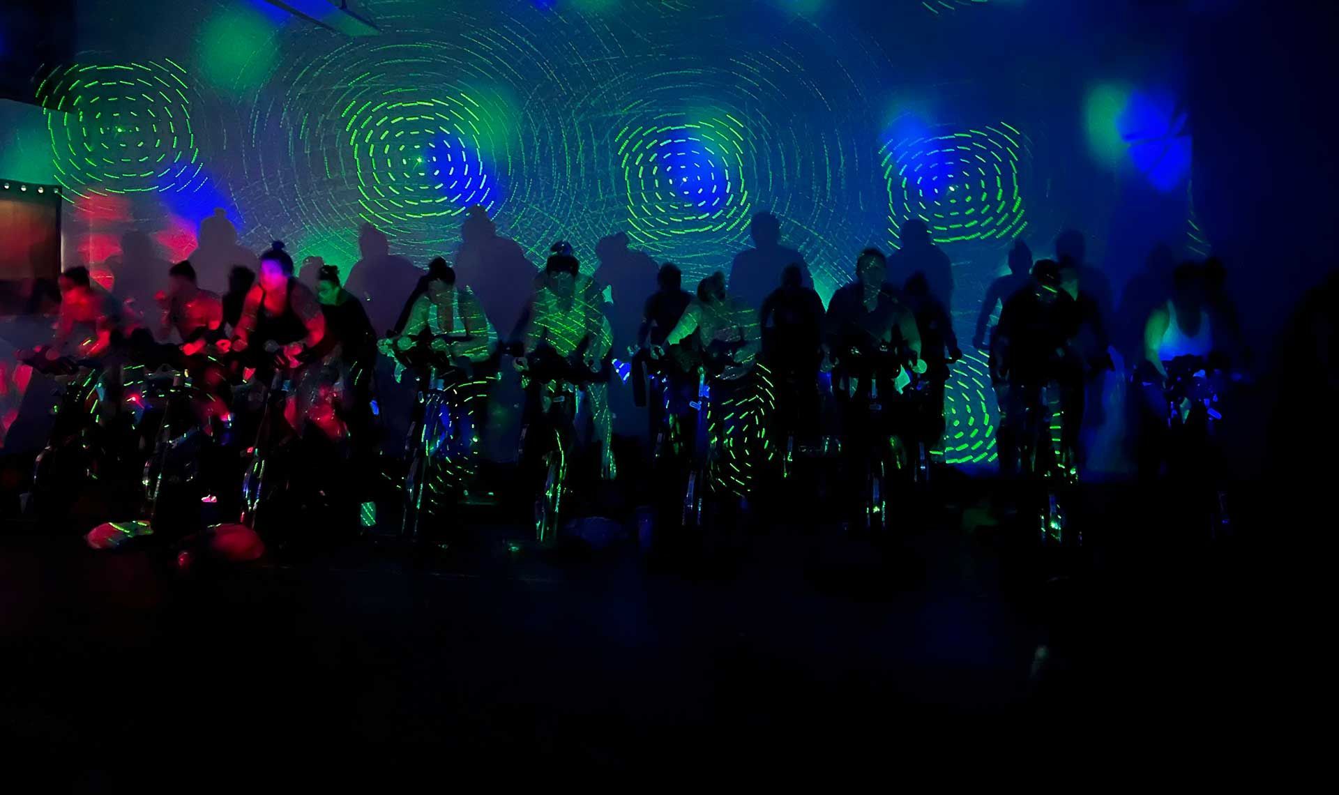 A group of people are riding indoor cycling bikes in a dark room